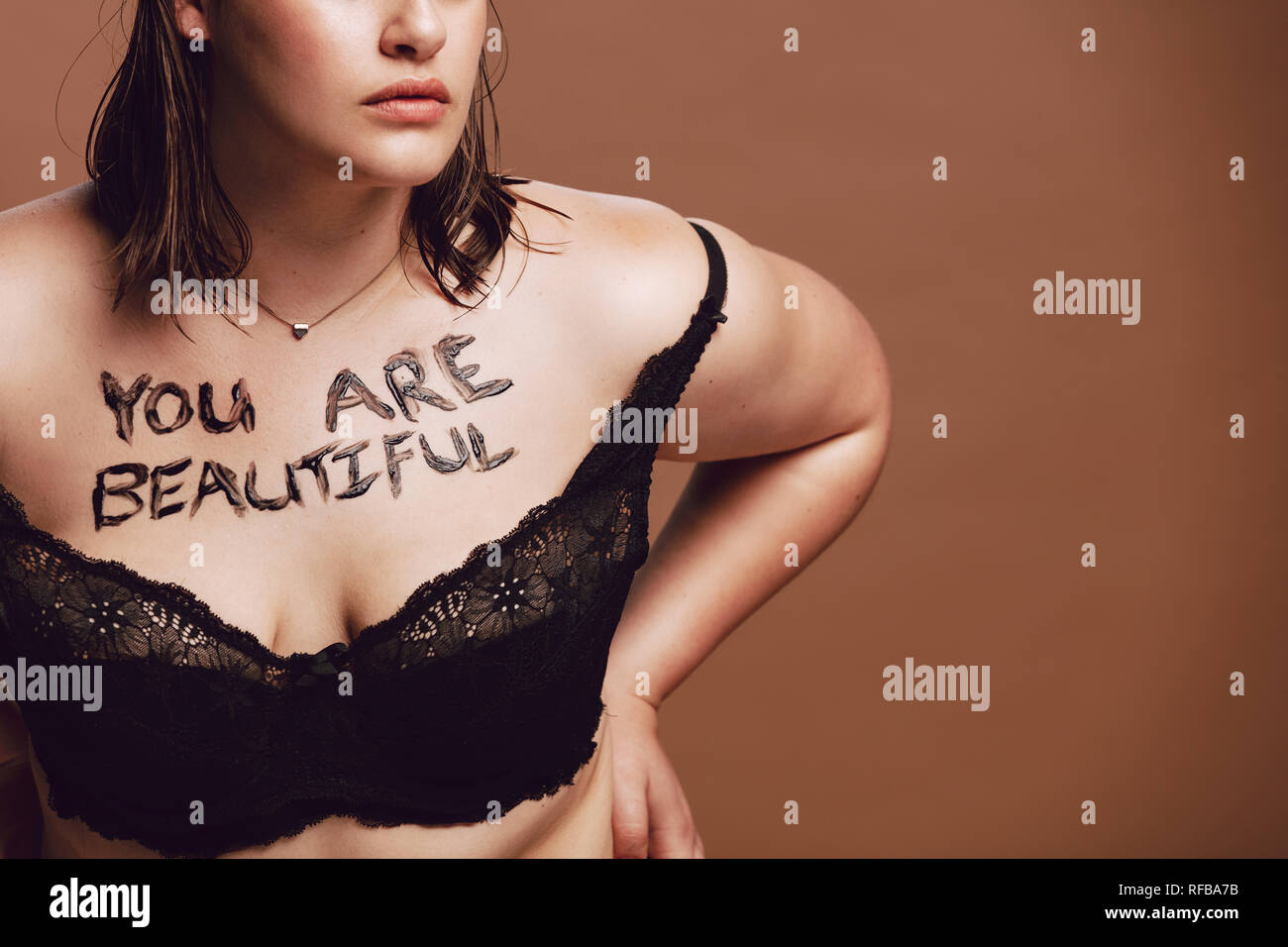 Cropped shot of woman surdimensionnée en soutien-gorge noir. Femme avec "Vous êtes belles", écrit sur son corps contre fond brun Banque D'Images