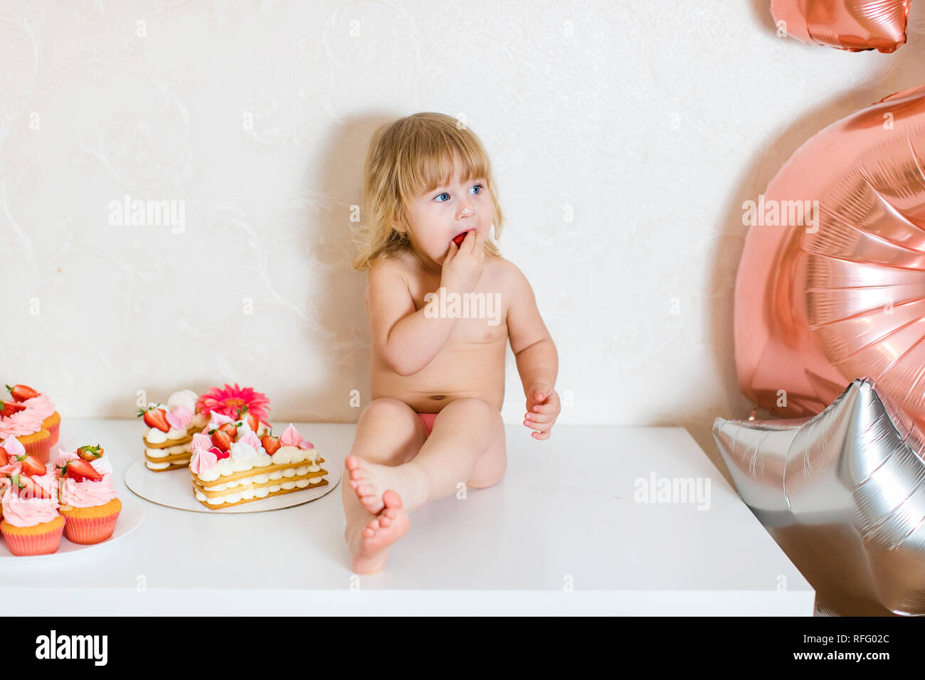 Petite blonde fille bébé deux ans en pantalon rose blanc assis sur la table près de son gâteau d'anniversaire et autre bonbons rose sur la table. Banque D'Images