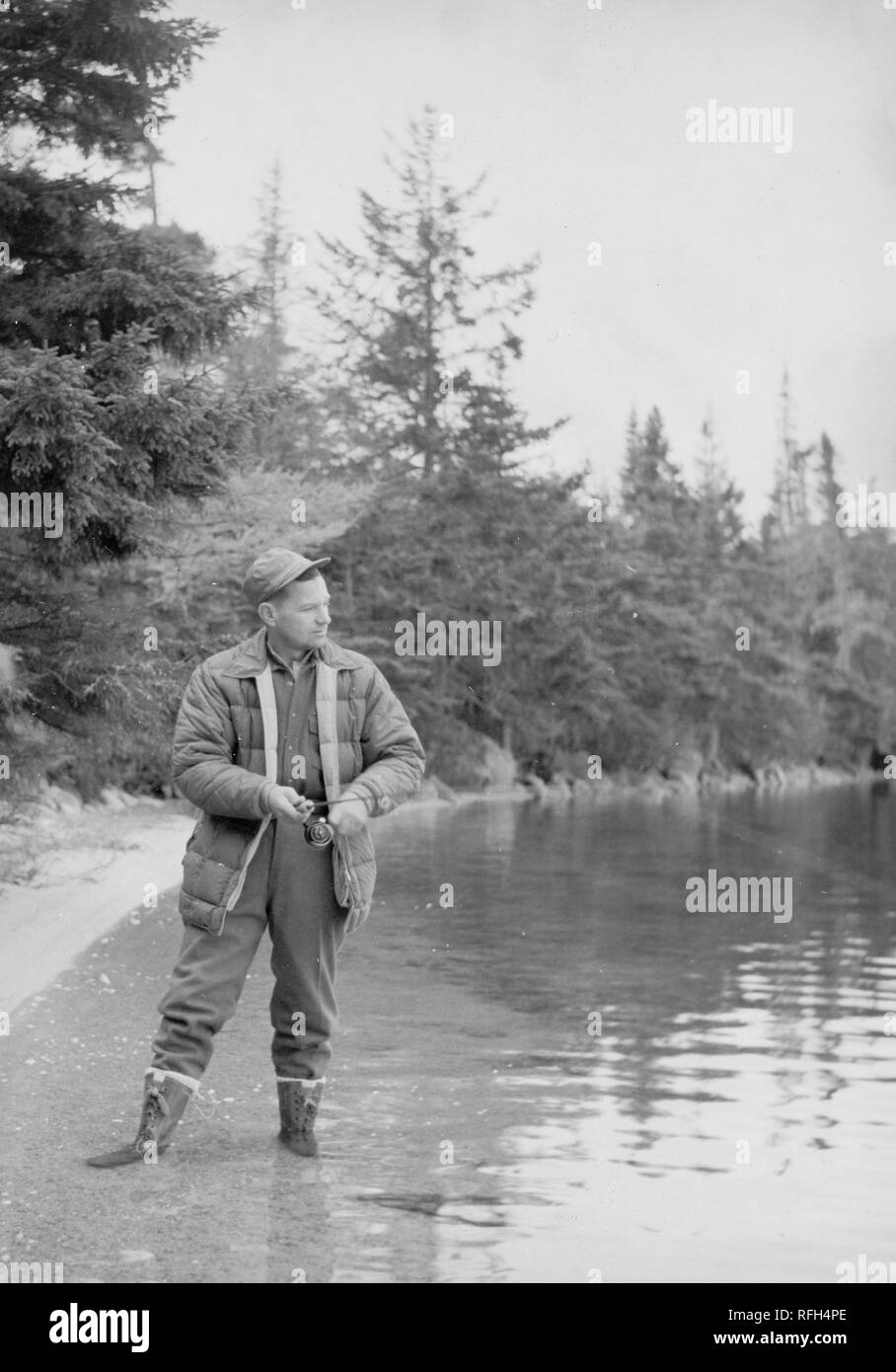 Photographie en noir et blanc d'un homme d'âge moyen, portant une chemise, salopettes, bottes, et le chapeau, illustré à la vue pleine longueur, debout dans un plan d'eau avec ses bottes à la cheville submergées, avec la tête tournée dans le profil de regarder vers la ligne d'une canne à pêche qu'il a avec les deux mains, avec des arbres et du feuillage en arrière-plan, photographié au cours d'un voyage de chasse situé en Alaska, USA, 1955. () Banque D'Images
