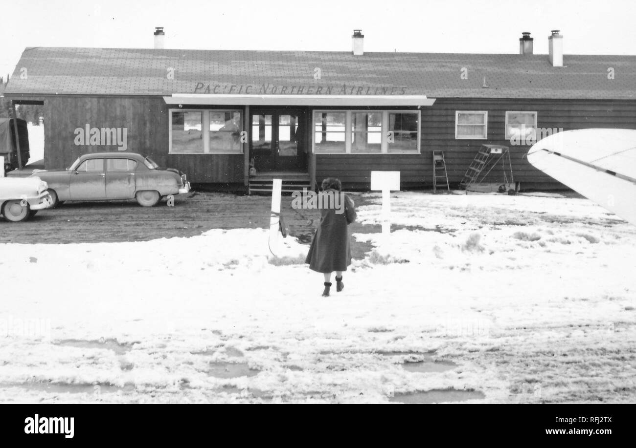 Photographie en noir et blanc d'une femme, tourné par derrière, en vue pleine longueur, portant une longue robe sombre, garni de fourrure et des bottes, marchant sur sol couvert de neige vers la porte ouverte d'un sol en bois, un seul niveau terminal de l'aéroport marqué 'Pacific Northern Airlines, ' avec les ailes d'un avion visible à droite au premier plan, photographié au cours d'un voyage de chasse, de pêche situé dans l'Alaska, 1955. () Banque D'Images