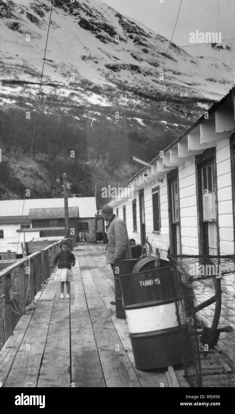 Photographie en noir et blanc d'une petite fille aux cheveux bruns, portant une veste sur une jupe ou une robe, debout sur une jetée en bois, face à la caméra en vue de pleine longueur, avec un homme portant une chemise et le chapeau, visible de profil à droite, metal barils et un fil piège de pêche s'appuyer contre un bâtiment en bois à droite au premier plan, et les pentes couvertes de neige sont visibles en arrière-plan ; photographié au cours d'un voyage de chasse, de pêche situé dans l'Alaska, 1955. () Banque D'Images