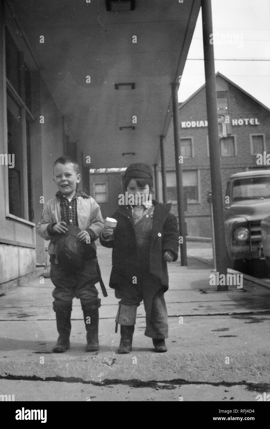 Photographie en noir et blanc de deux petits garçons, souriant, debout ensemble en vue de pleine longueur sur un trottoir au centre-ville de Kodiak, Alaska, les deux portant des manteaux chauds et des bottes, l'un tenant une glace partiellement mangés-cornet de crème glacée et l'autre tenant son chapeau, avec un chariot vintage et un bâtiment avec le signe 'Hôtel Kodiak' visible dans l'arrière-plan, photographié au cours d'un voyage de chasse, de pêche situé dans l'Alaska, USA, 1955. () Banque D'Images