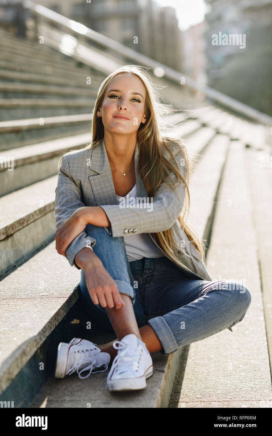 Belle jeune femme de race blanche en contexte urbain. Fille blonde avec une veste et un jean bleu assis sur des escaliers. Banque D'Images