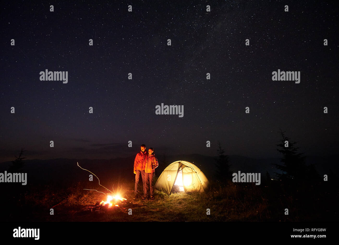 Couple heureux randonneurs boy and Girl standing in front of tente bien éclairé par le feu de joie sous un ciel étoilé, profitant de nuit tranquille dans les montagnes. Touris Banque D'Images