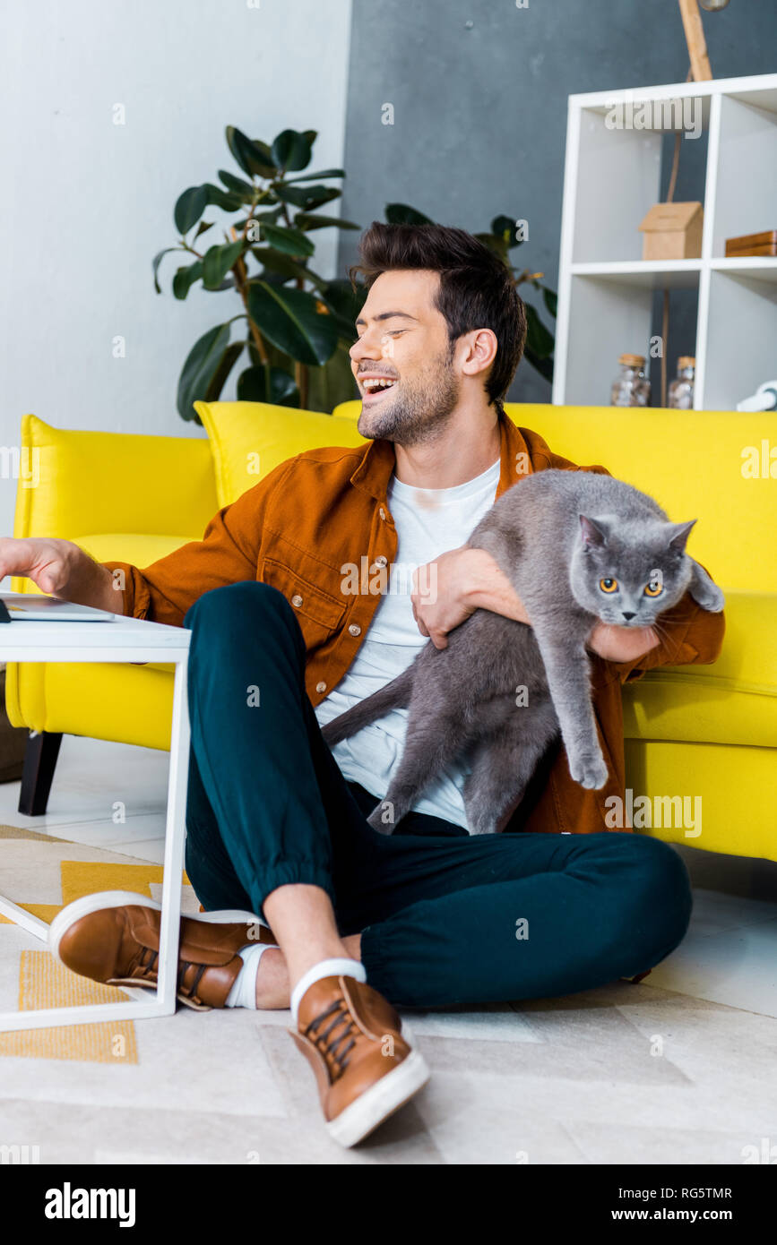 Les jeunes laughing man using laptop while sitting on floor with chat gris Banque D'Images