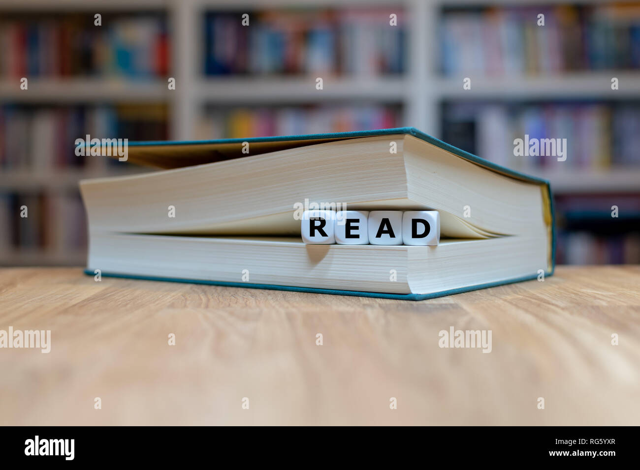 Dés dans un livre forment le mot 'read'. Livre est couchée sur un bureau en bois en face d'une étagère. Banque D'Images