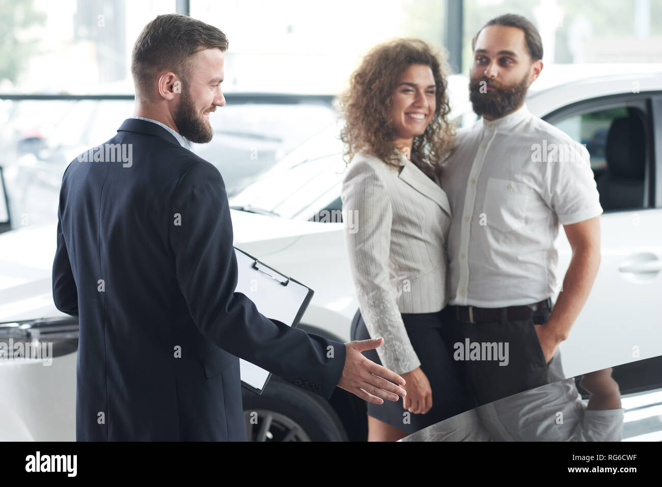 Couple communiquant avec manager dans l'agence de voiture. Voiture de porter en veste bleu foncé montrant avec la main des véhicules. Belle femme et homme barbu L'observation d'automobiles. Banque D'Images