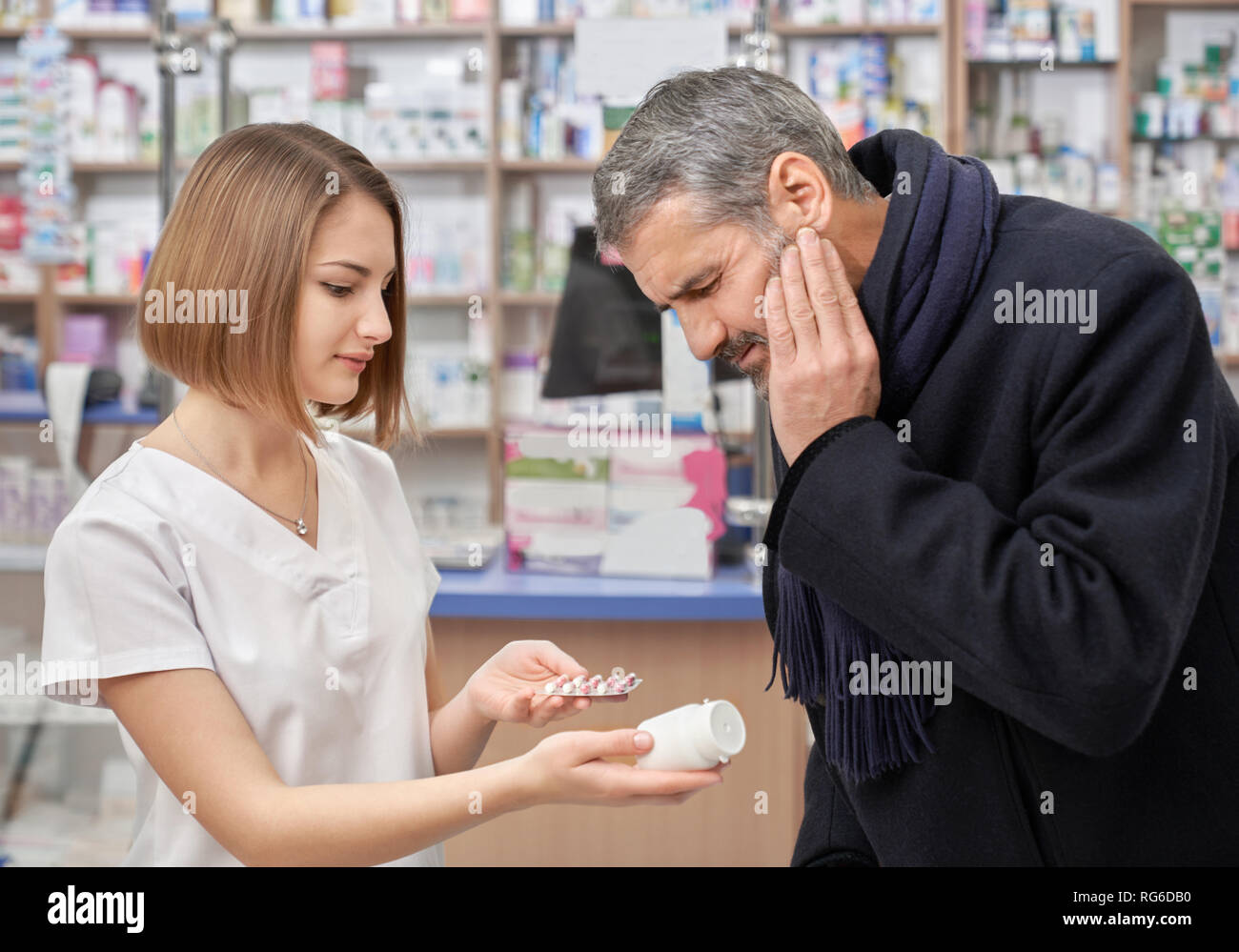 Mal de dent senior consulting avec votre pharmacien. Jolie femme en uniforme blanc offre des médicaments, holding pill bottle et blister en mains. L'homme aux yeux clos fronçant douloureusement. Banque D'Images