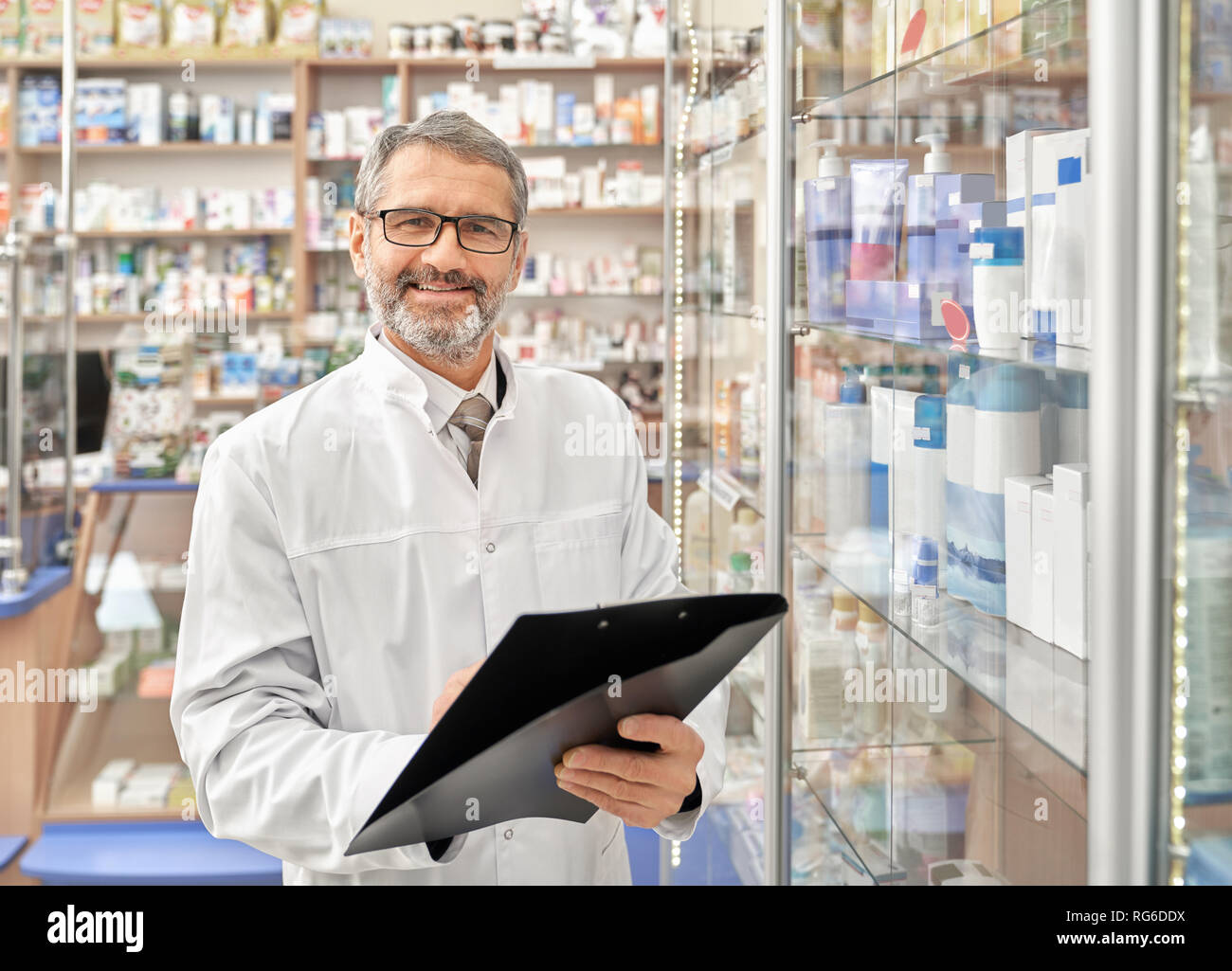 Young man smiling, pose, debout près des étagères de pharmacie avec des médicaments du dossier. Beau pharmacien barbu avec les cheveux gris vêtu de blanc sarrau et lunettes. Banque D'Images