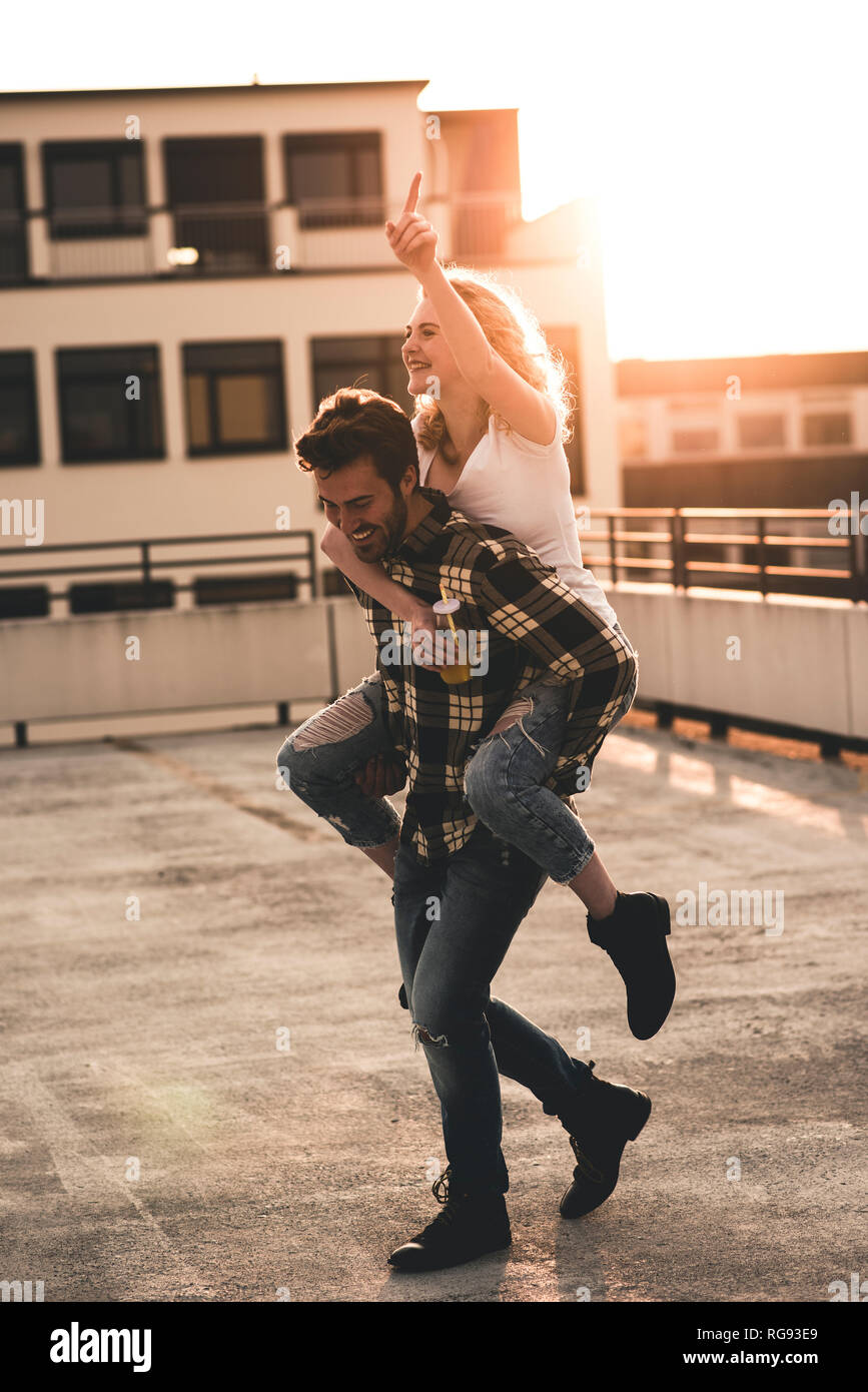 Happy young man giving his girlfriend piggyback ride sur un toit-terrasse au coucher du soleil Banque D'Images
