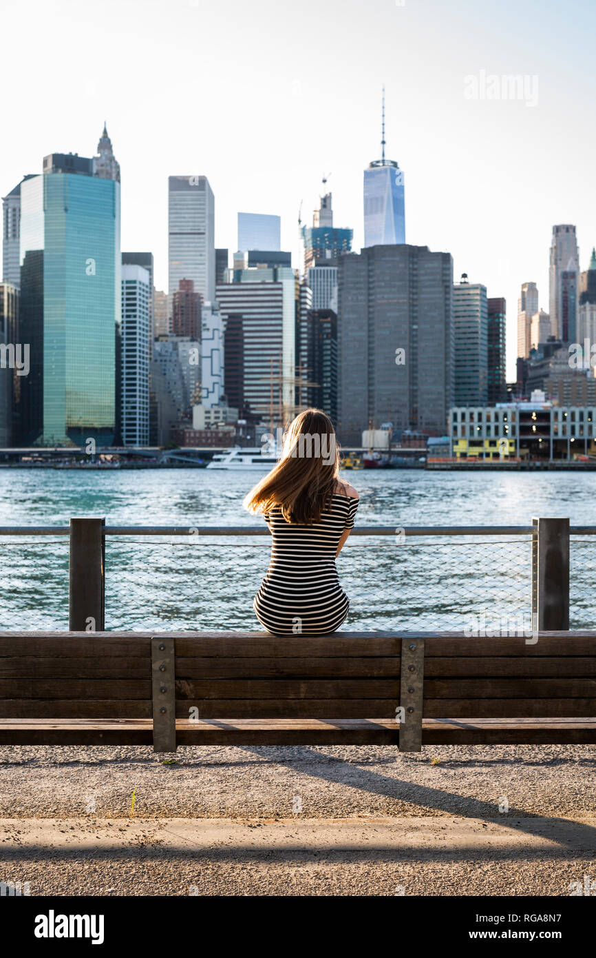 USA, New York, Brooklyn, vue arrière de femme assis sur un banc en face du fleuve et les toits de Manhattan Banque D'Images