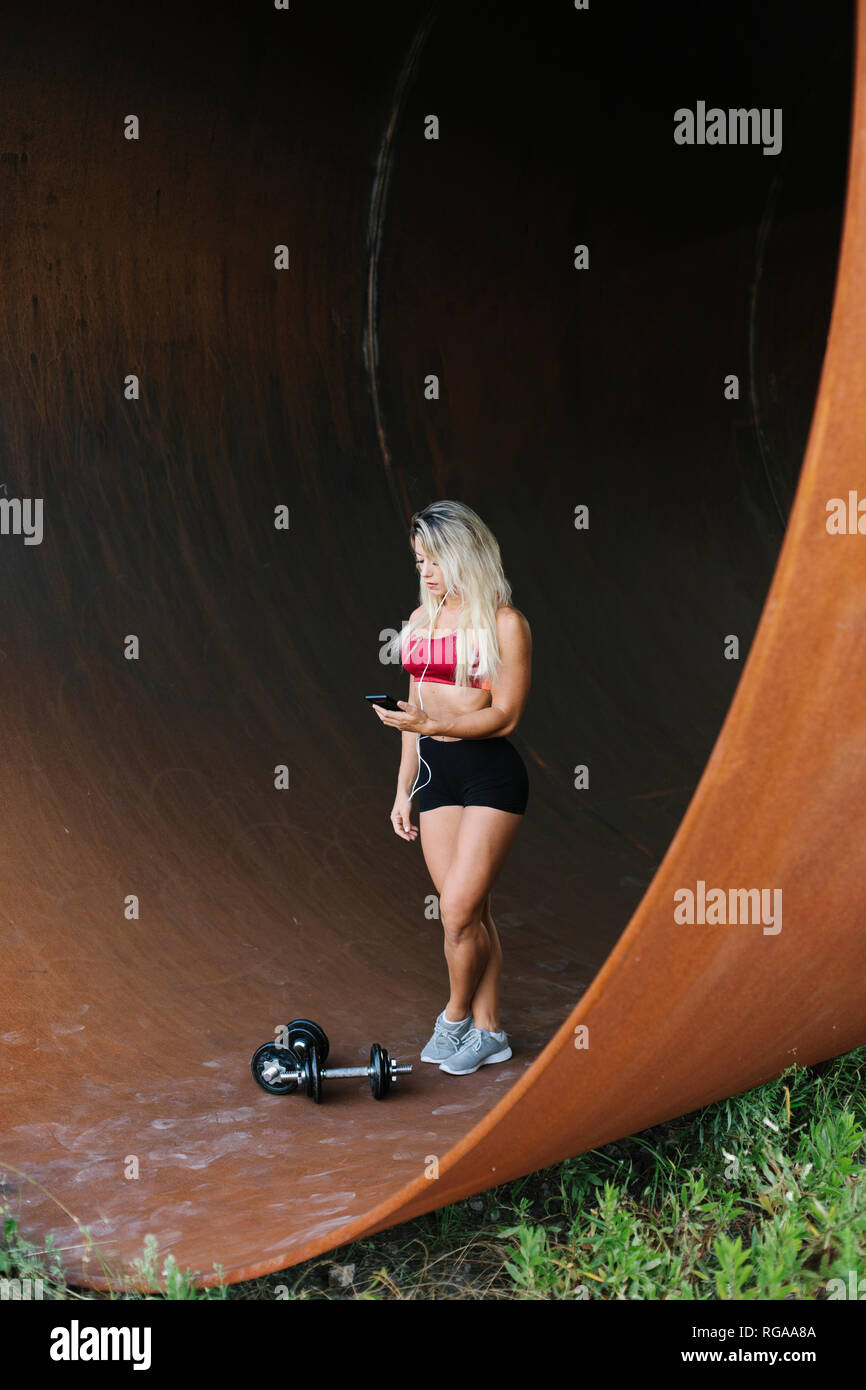 Athletic femme debout à l'intérieur d'un tube avec haltères et écouteurs looking at cell phone Banque D'Images