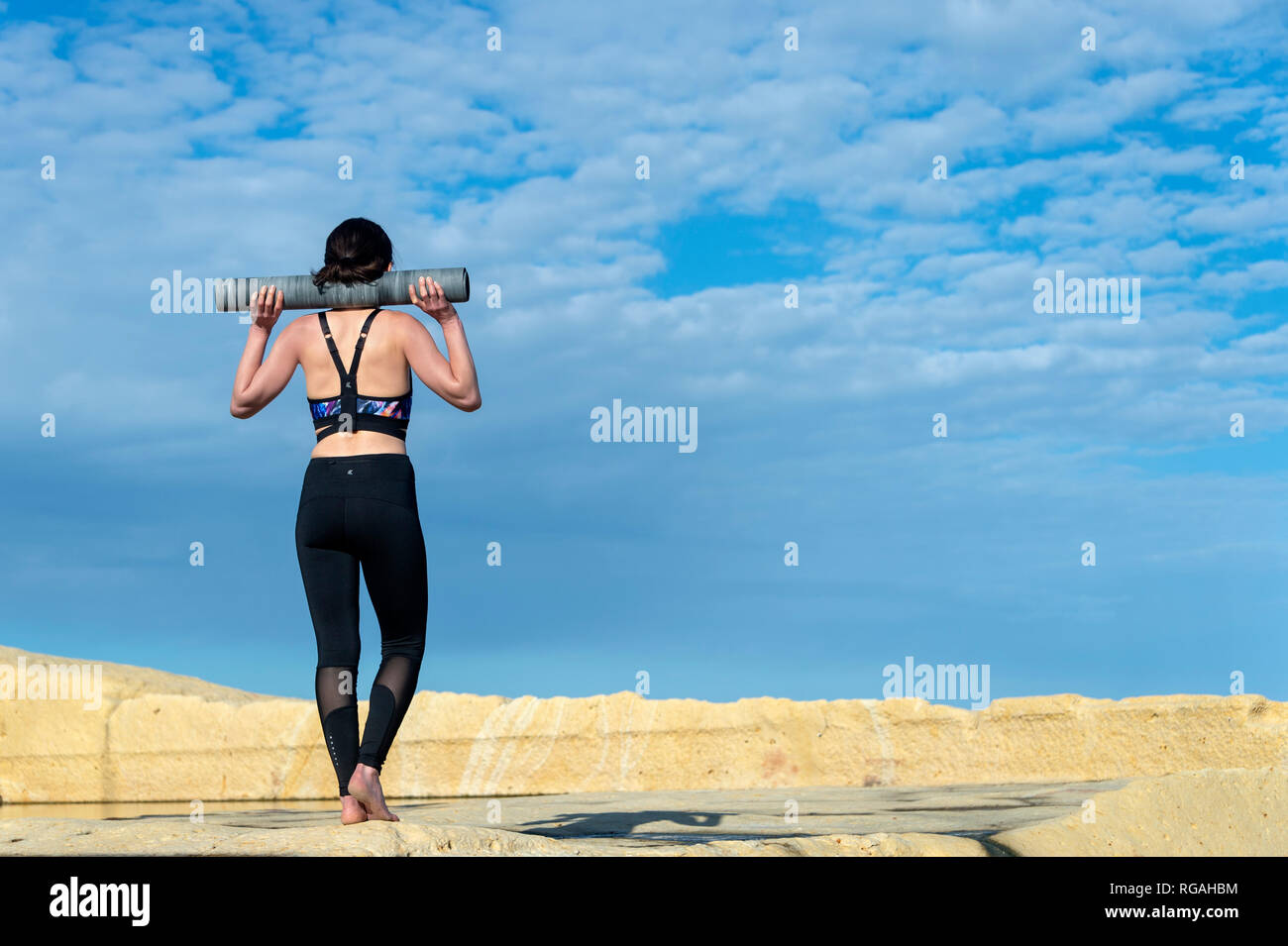 Woman wearing sportswear transportant un tapis de yoga sur ses épaules à l'extérieur au soleil avec ciel bleu Banque D'Images