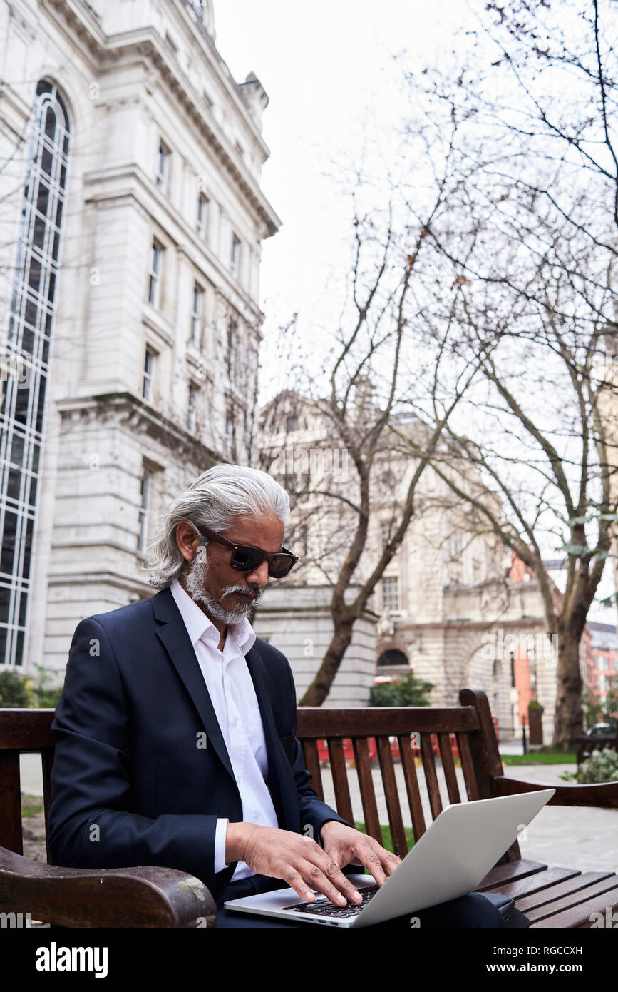 UK, Londres, senior businessman sitting on bench outdoors working on laptop Banque D'Images