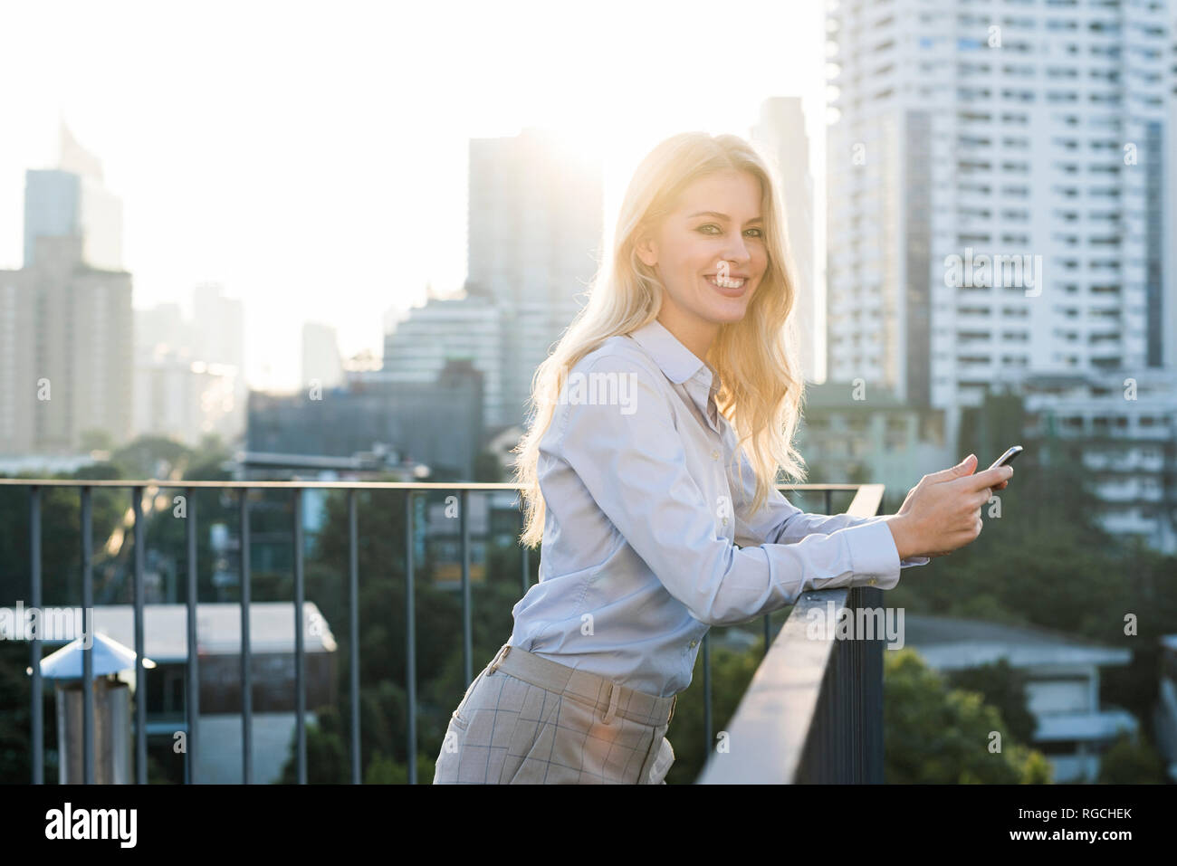 Blonde smiling business woman holding smartphone sur main courante sur les toits de la ville Banque D'Images