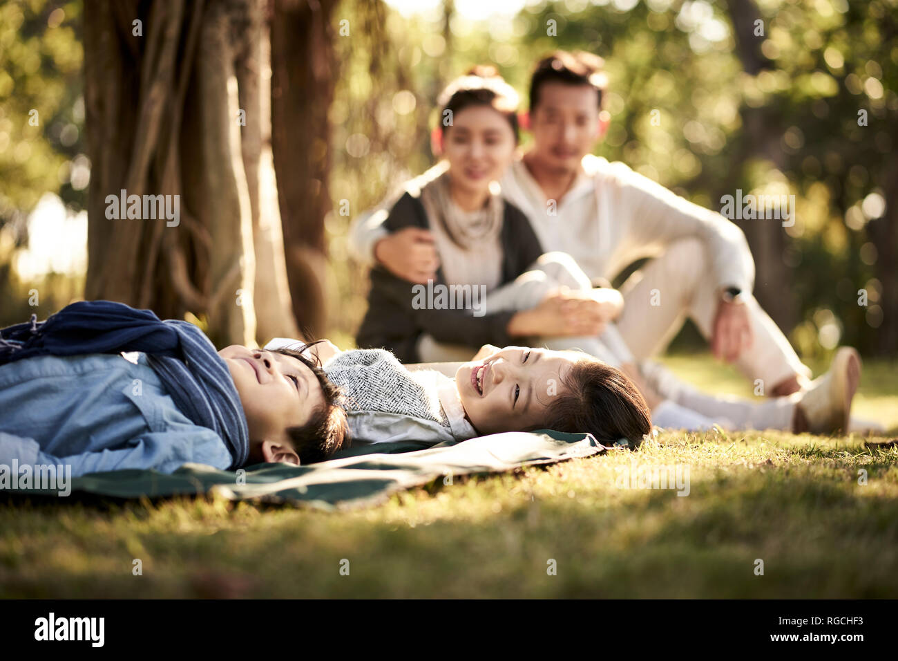 Deux enfants asiatiques little boy and girl having fun lying on grass avec les parents assis à regarder en arrière-plan. Banque D'Images