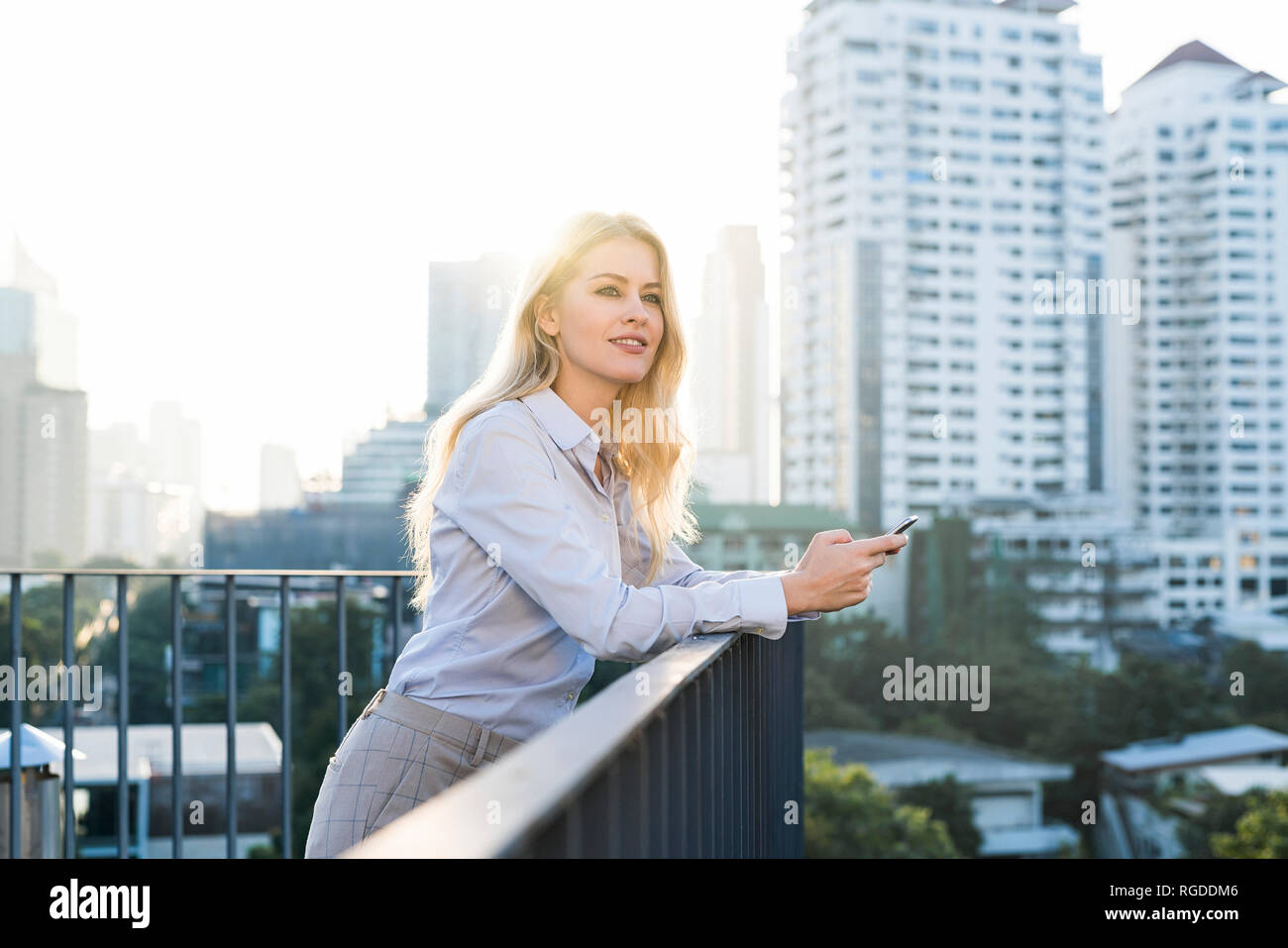 Blonde smiling business woman holding smartphone sur main courante sur les toits de la ville Banque D'Images