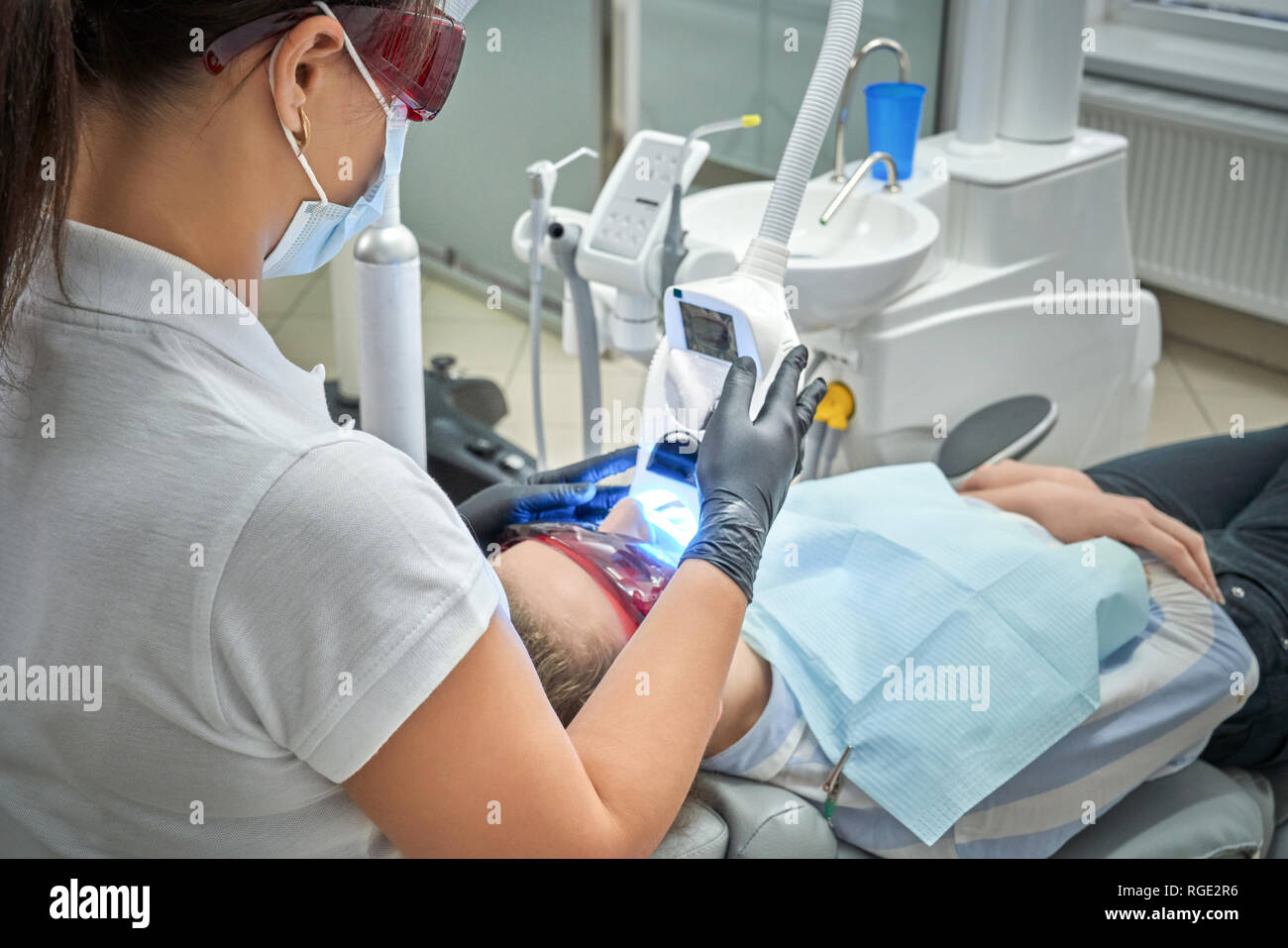 Vue de l'arrière du dentiste professionnel en train de faire de blanchiment des dents pour femme. Couché sur Client fauteuil dentaire en lunettes de protection tout en faisant médecin procédure avec équipement moderne. Banque D'Images