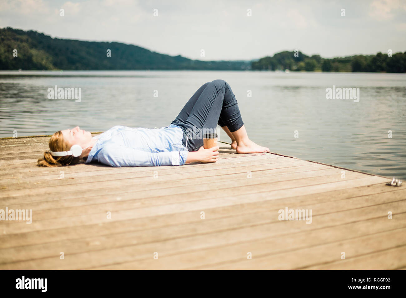 Woman lying on jetée à un lac avec des écouteurs et du café à emporter Banque D'Images Woman lying on jetée à un lac avec des écouteurs et du café à emporter Banque D'Images