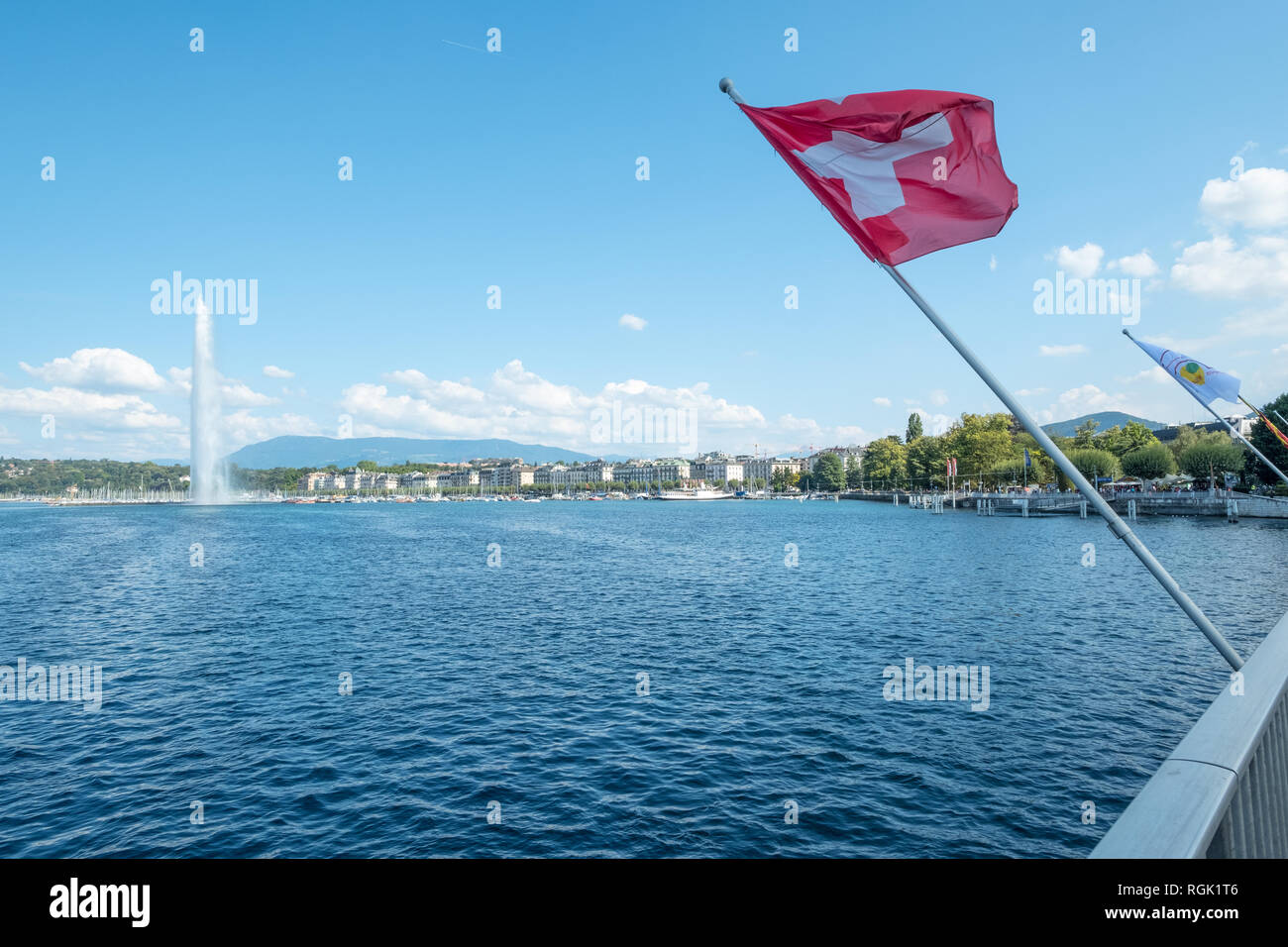 La photo en diagonale, en se penchant sur le lac de Genève, d'un drapeau rouge avec croix blanche emblématique survole un contexte, y compris le secteur riverain de Genève un Banque D'Images
