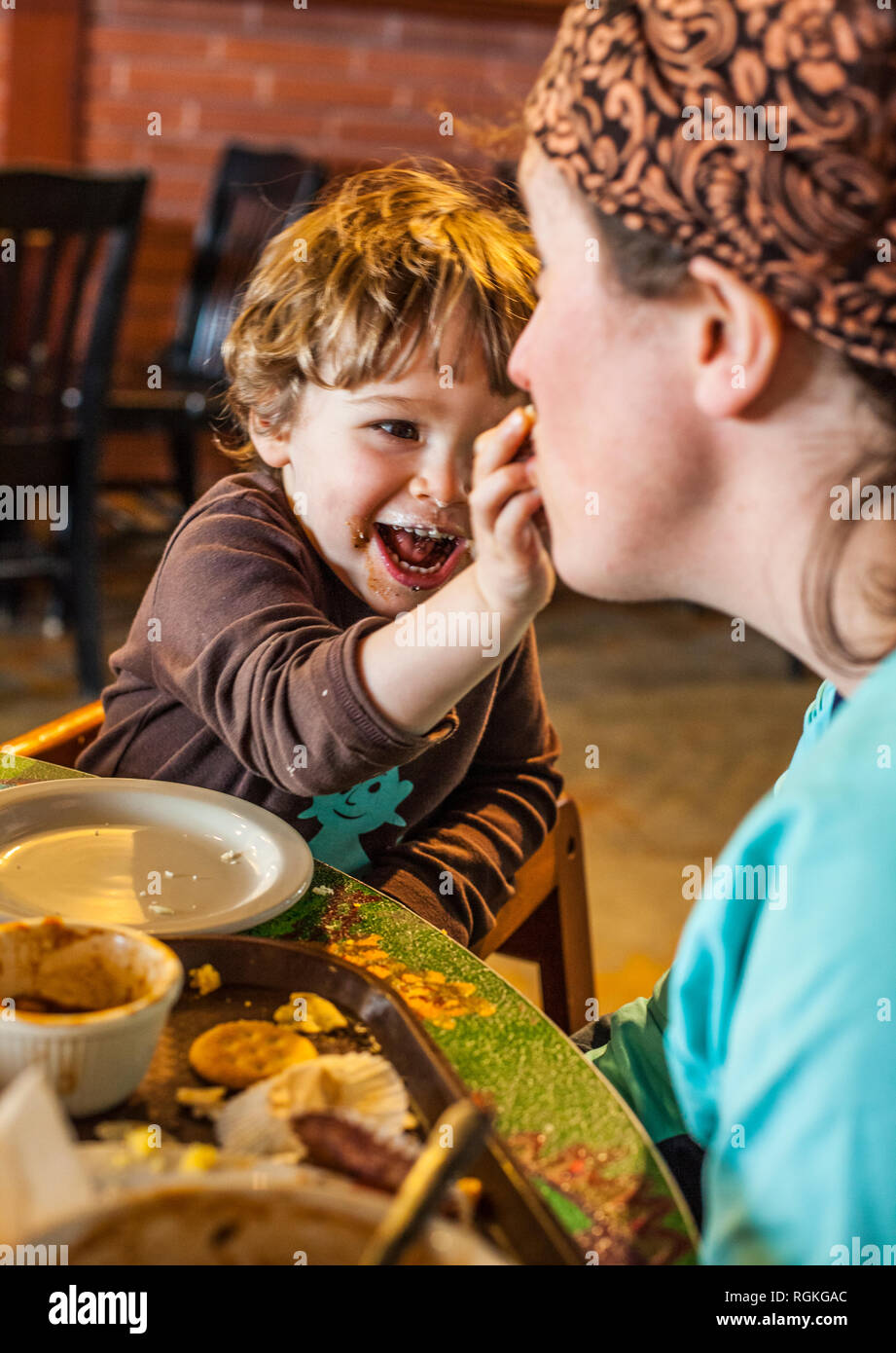 Un sale face toddler mettre la nourriture dans sa bouche et les mères de rire. Banque D'Images