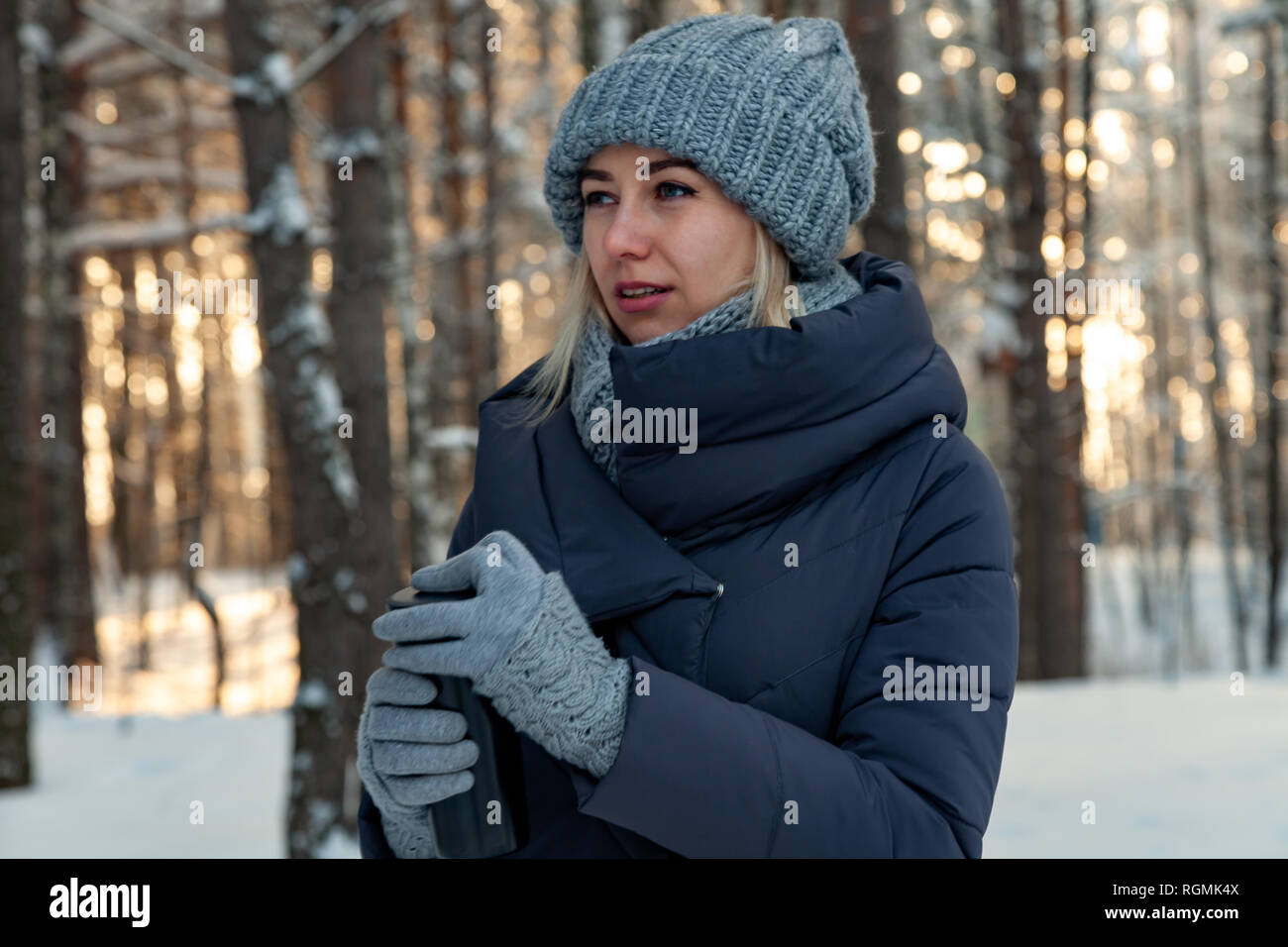 Belle jeune blonde en tricot de bonnet, écharpe et gants boit du café ou du  thé dans la forêt couverte de neige d'hiver au coucher du soleil de la  journée contre l'arrière