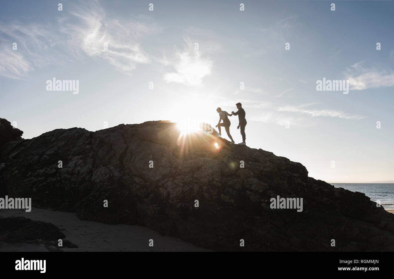 France, Bretagne, jeune couple d'escalade sur un rocher sur la plage au coucher du soleil Banque D'Images