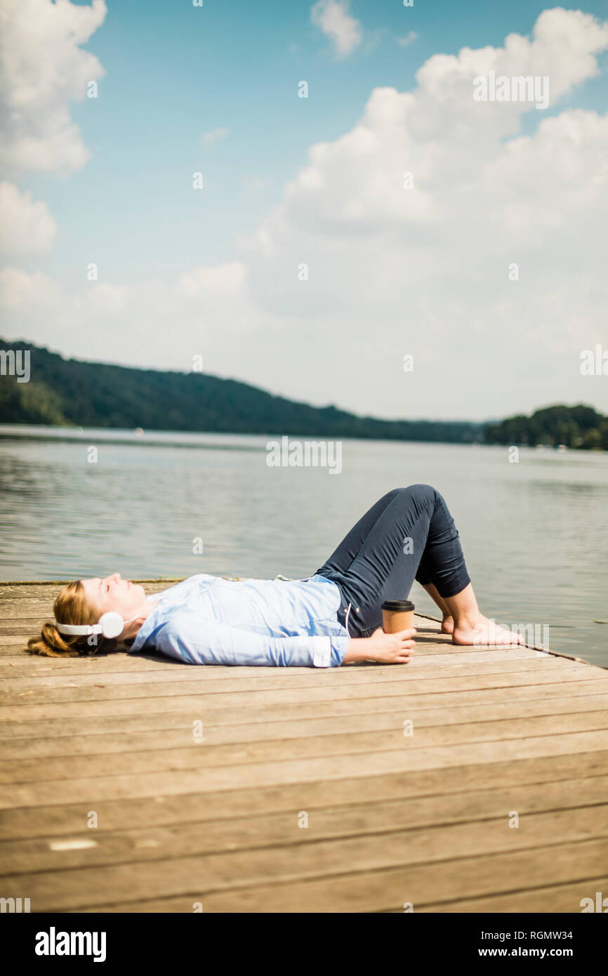 Woman lying on jetée à un lac avec des écouteurs et du café à emporter Banque D'Images Woman lying on jetée à un lac avec des écouteurs et du café à emporter Banque D'Images