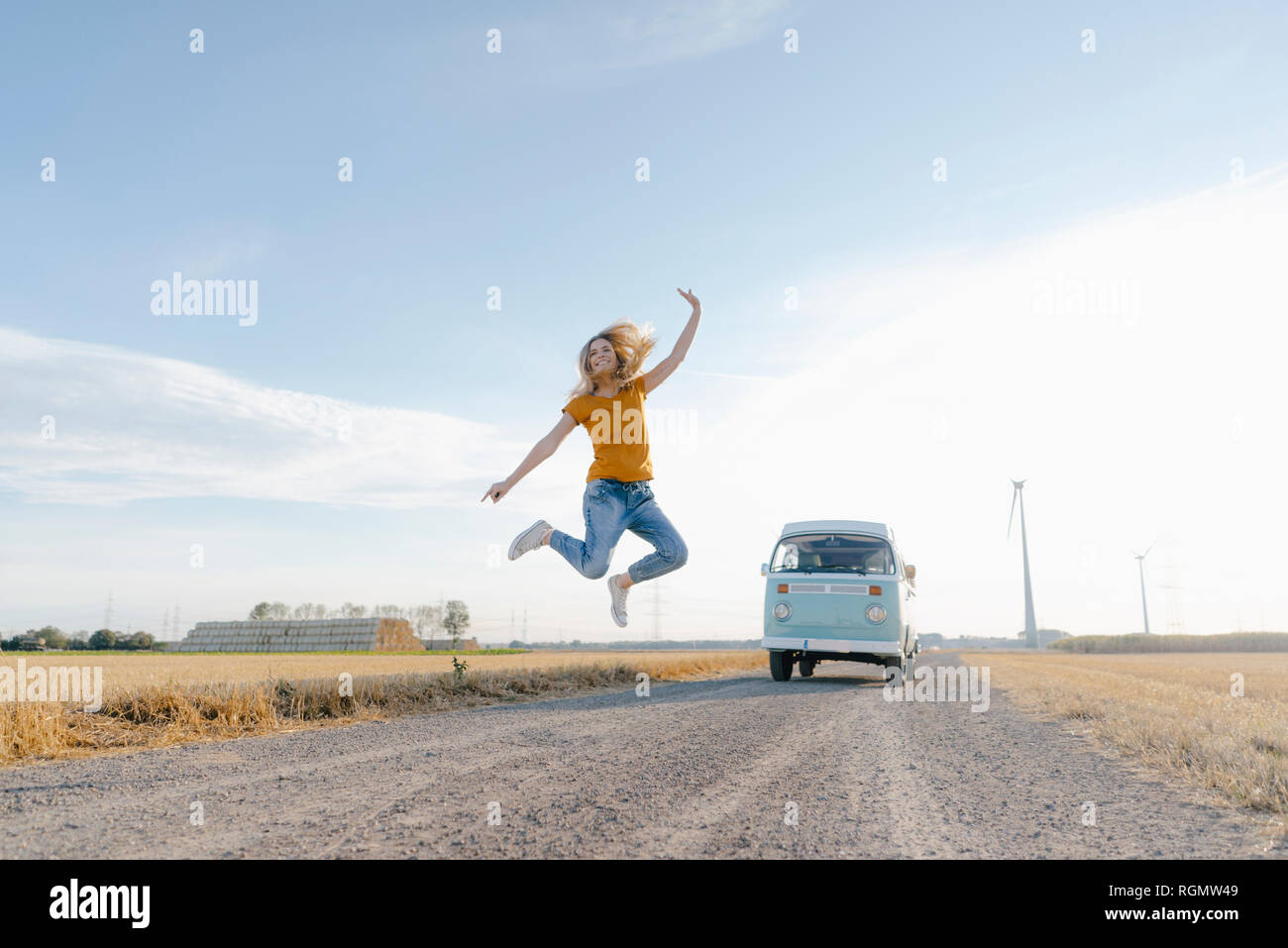 Jeune femme exubérante, sautant sur un chemin de terre à camper van in rural landscape Banque D'Images