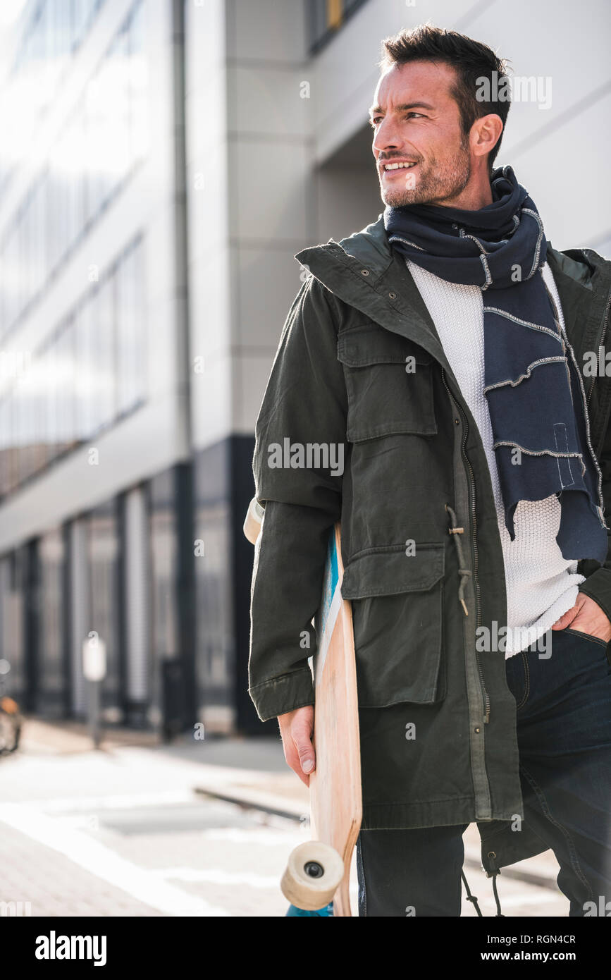 Homme mûr, portant manteau et écharpe, marcher dans la ville, faisant longboard Banque D'Images