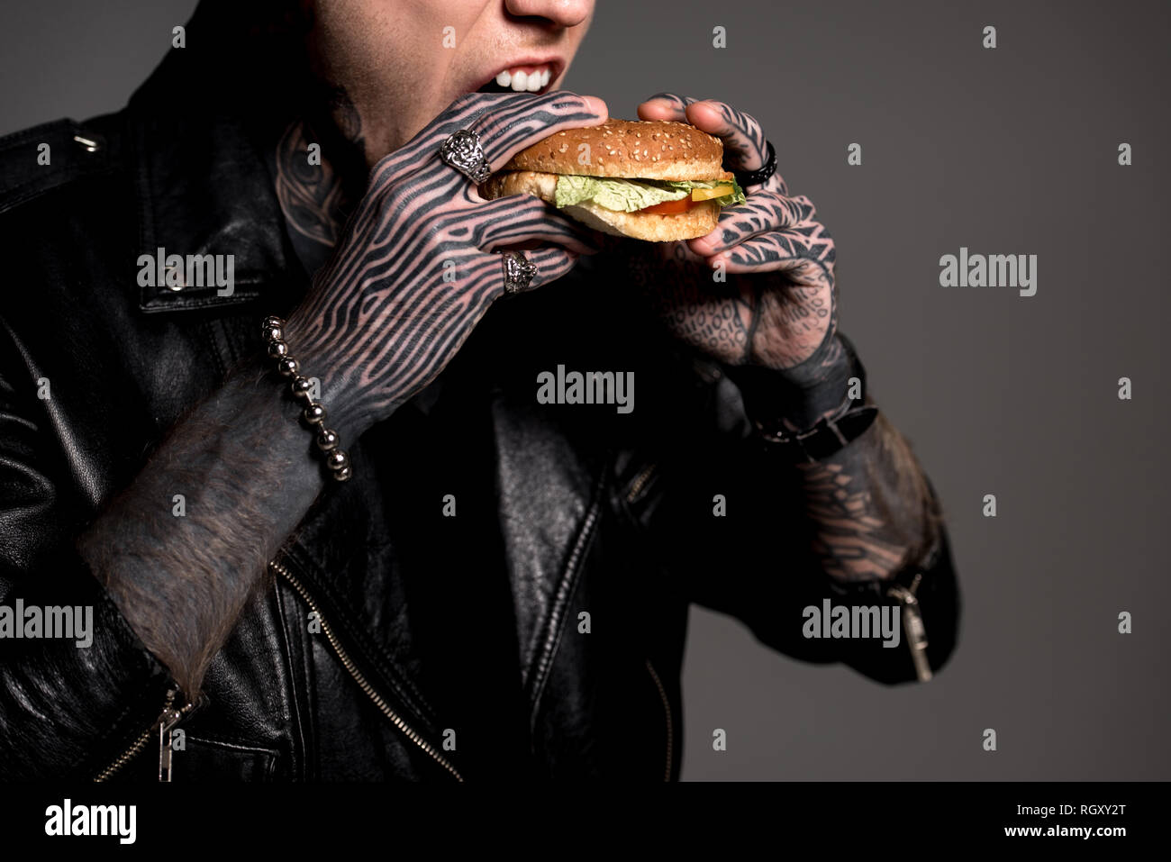 Cropped shot élégant de l'homme tatoué en blouson de cuir manger burger isolé sur gray Banque D'Images