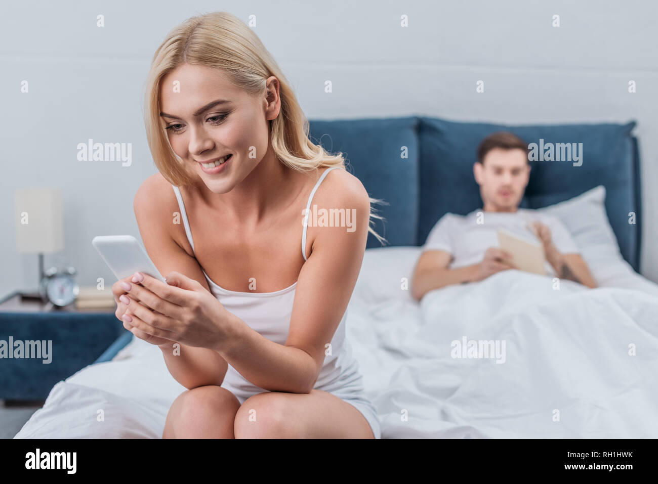 Smiling young woman sitting on bed and using smartphone alors que petit ami derrière reading book Banque D'Images