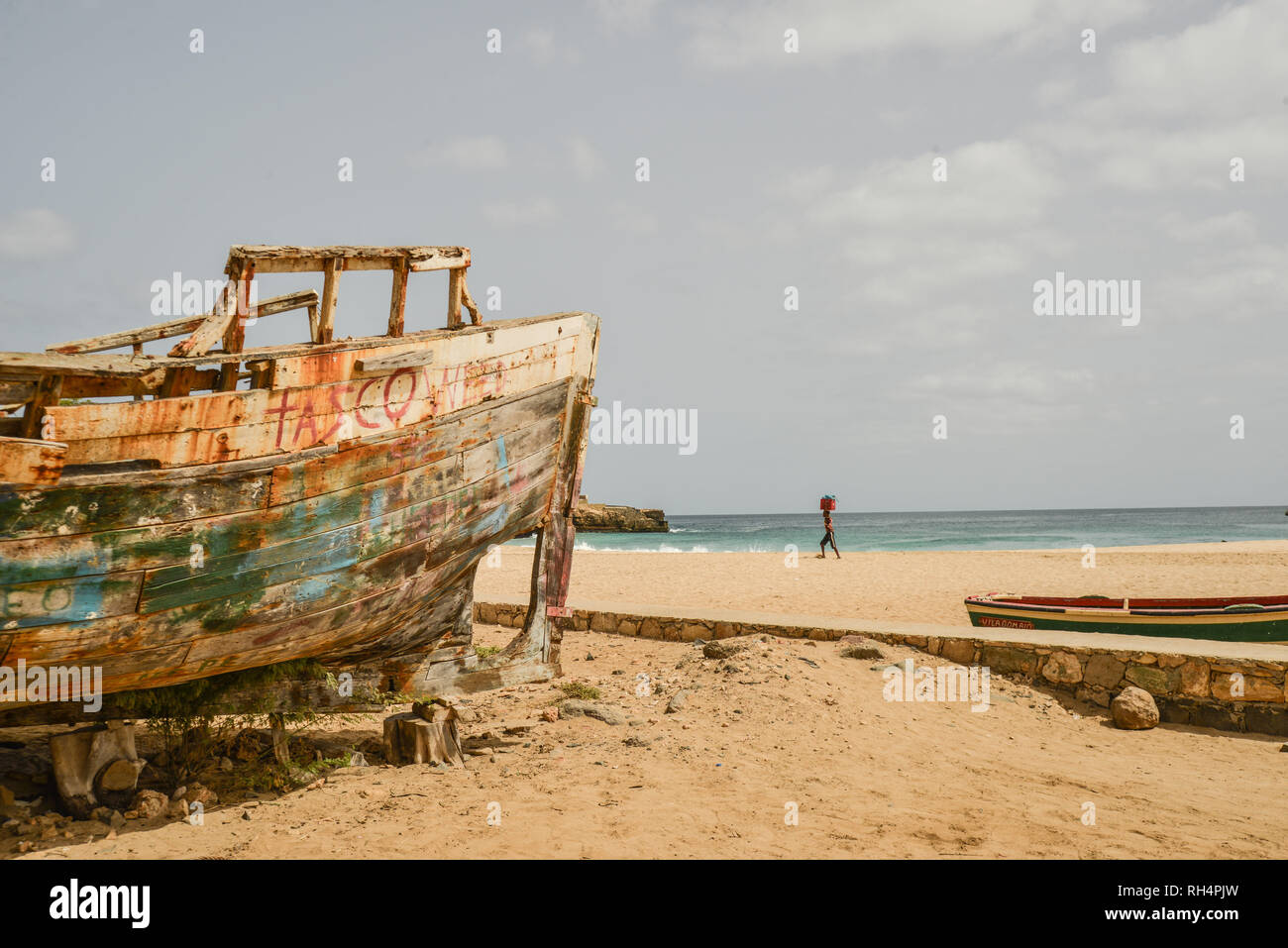 Le Cap Vert, l'île de Maio : barge abandonnée sur la plage de Porto Ingles (Vila do Maio) et jeune homme de la distance pour le transport de marchandises sur la tête Banque D'Images