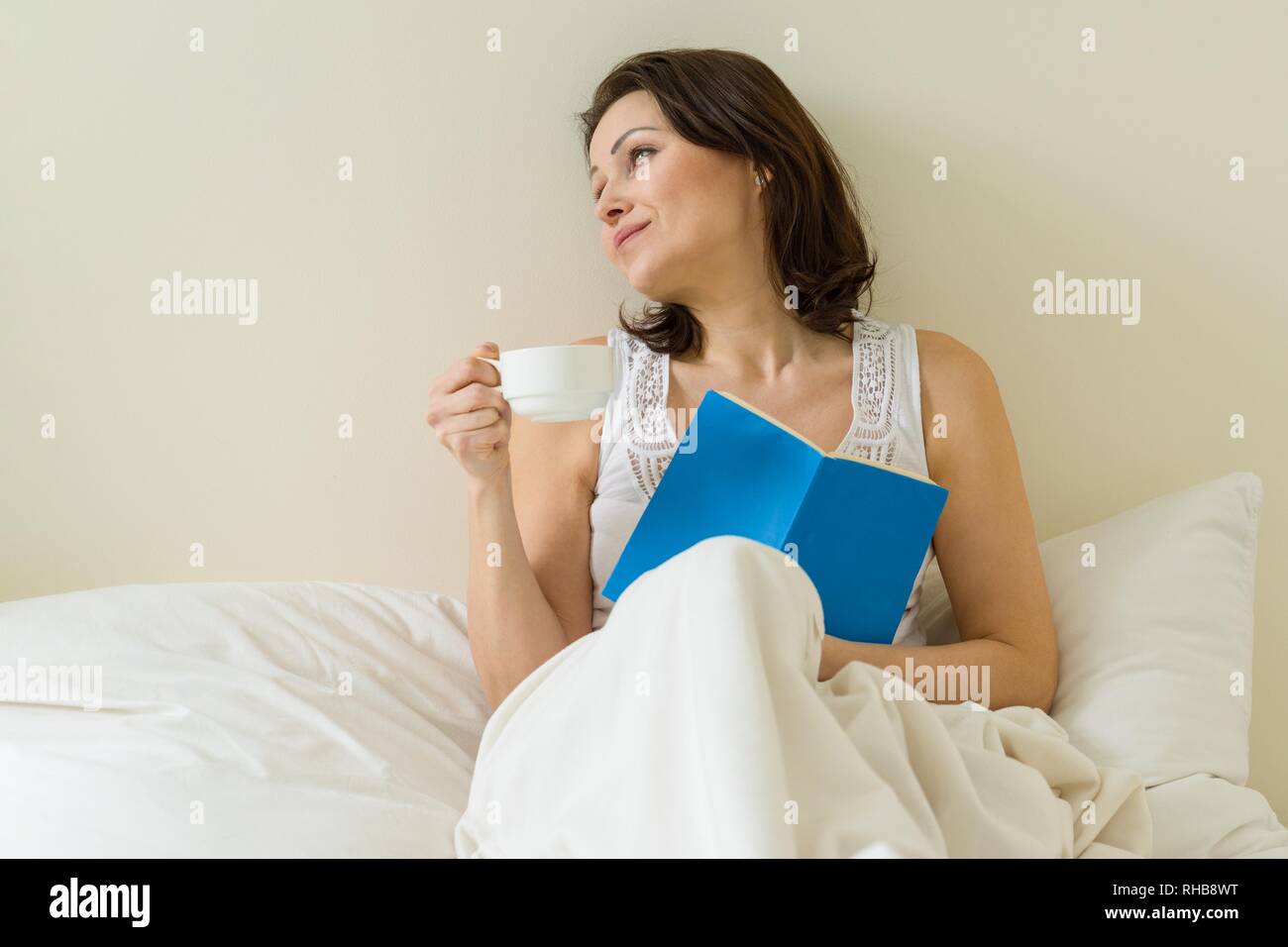 Mature Woman in bed reading book et de boire le café du matin. Banque D'Images
