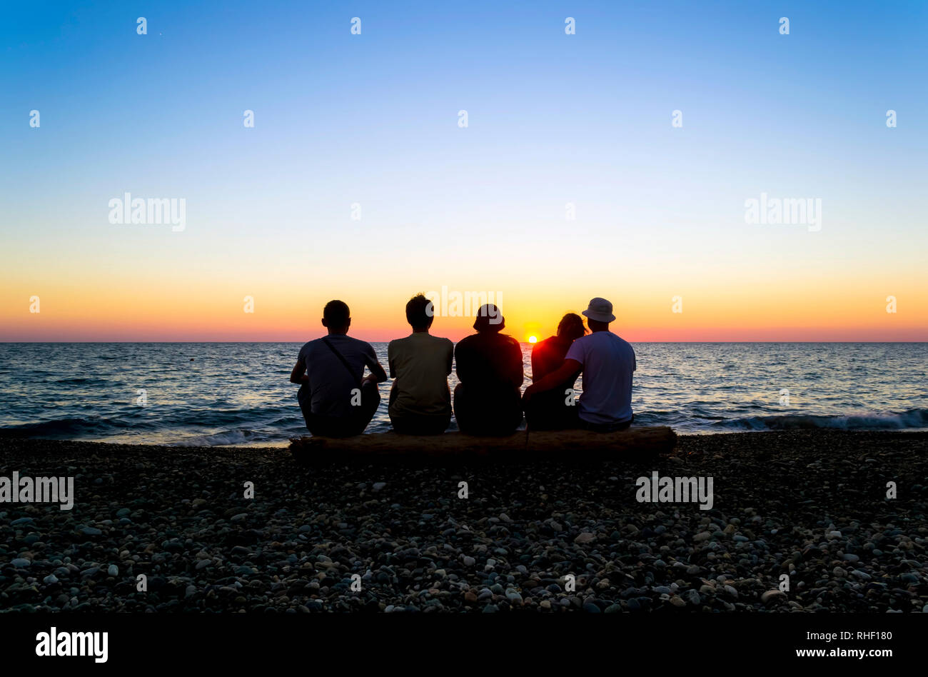 Un groupe de personnes assises sur la plage et regarder le coucher du soleil. Silhouettes de personnes contre le soleil. Ciel bleu clair. Plage en pierre. Jours fériés par la se Banque D'Images