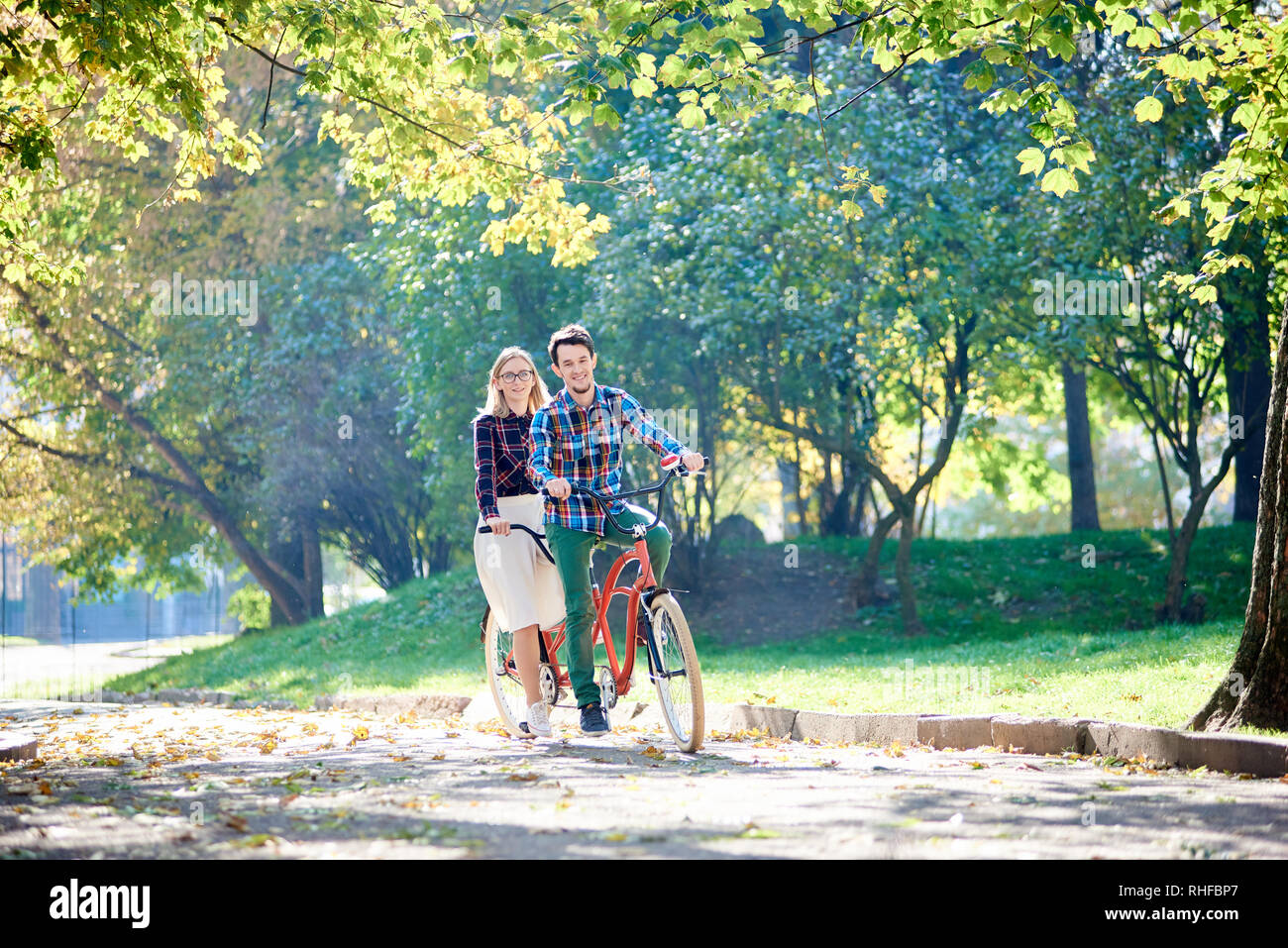 Jeune couple voyageur smiling actif, beau barbu et attractive blonde woman ensemble double tandem vélo le long chemin craquelé dans éclairée par le soleil du matin lumineux beau parc sous les arbres. Banque D'Images