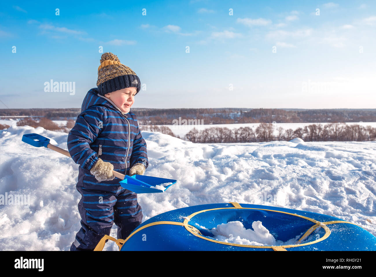 Un petit garçon enfant joue dans la neige en hiver. Espace libre pour le texte. En salopette bleue et un chapeau. Sur la Lame à neige porte tube. Banque D'Images