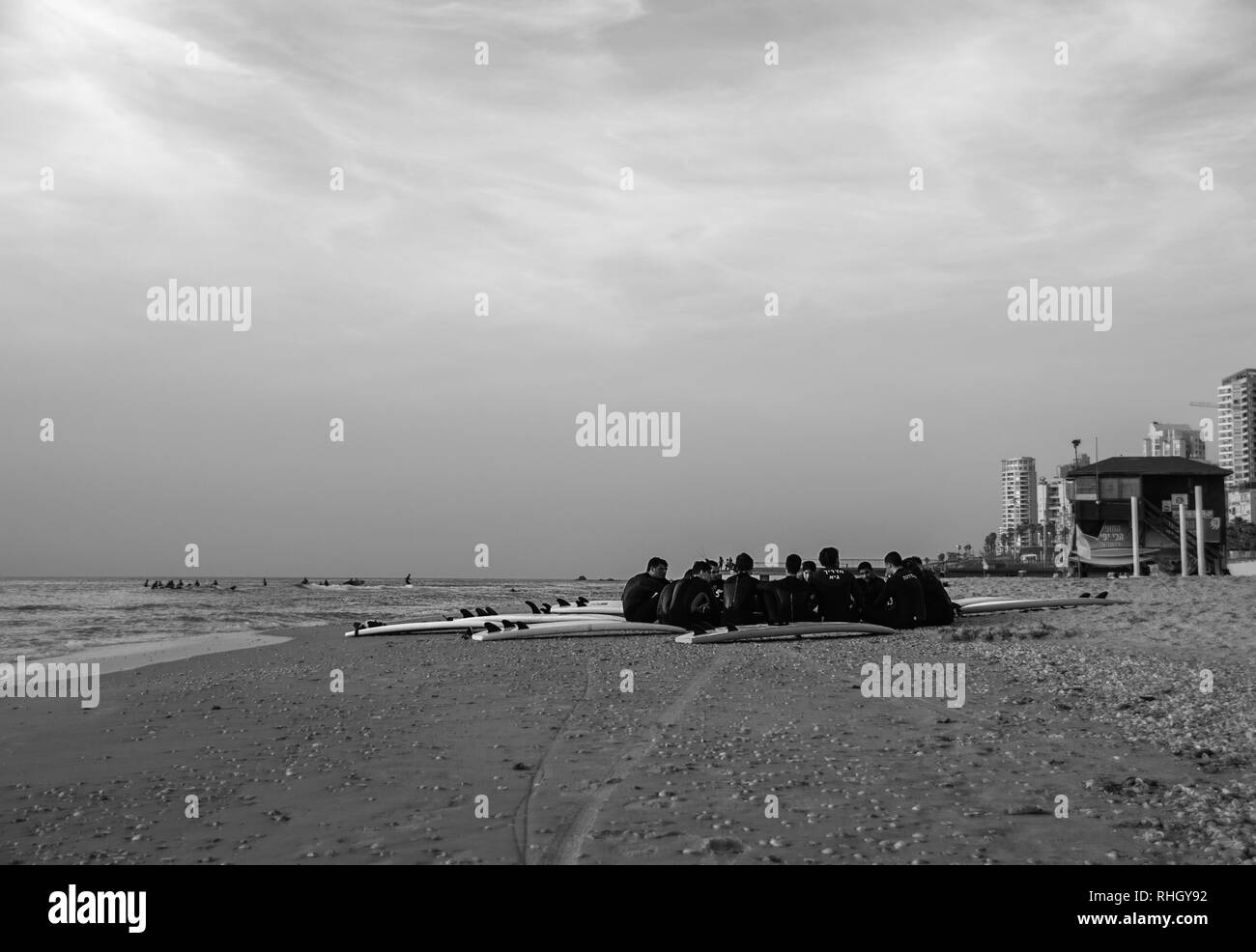 Cercle de surfeurs à combinaisons, assis sur une plage de sable fin par l'eau et d'un sauveteur shack, sous un ciel nuageux, en noir et blanc. Banque D'Images