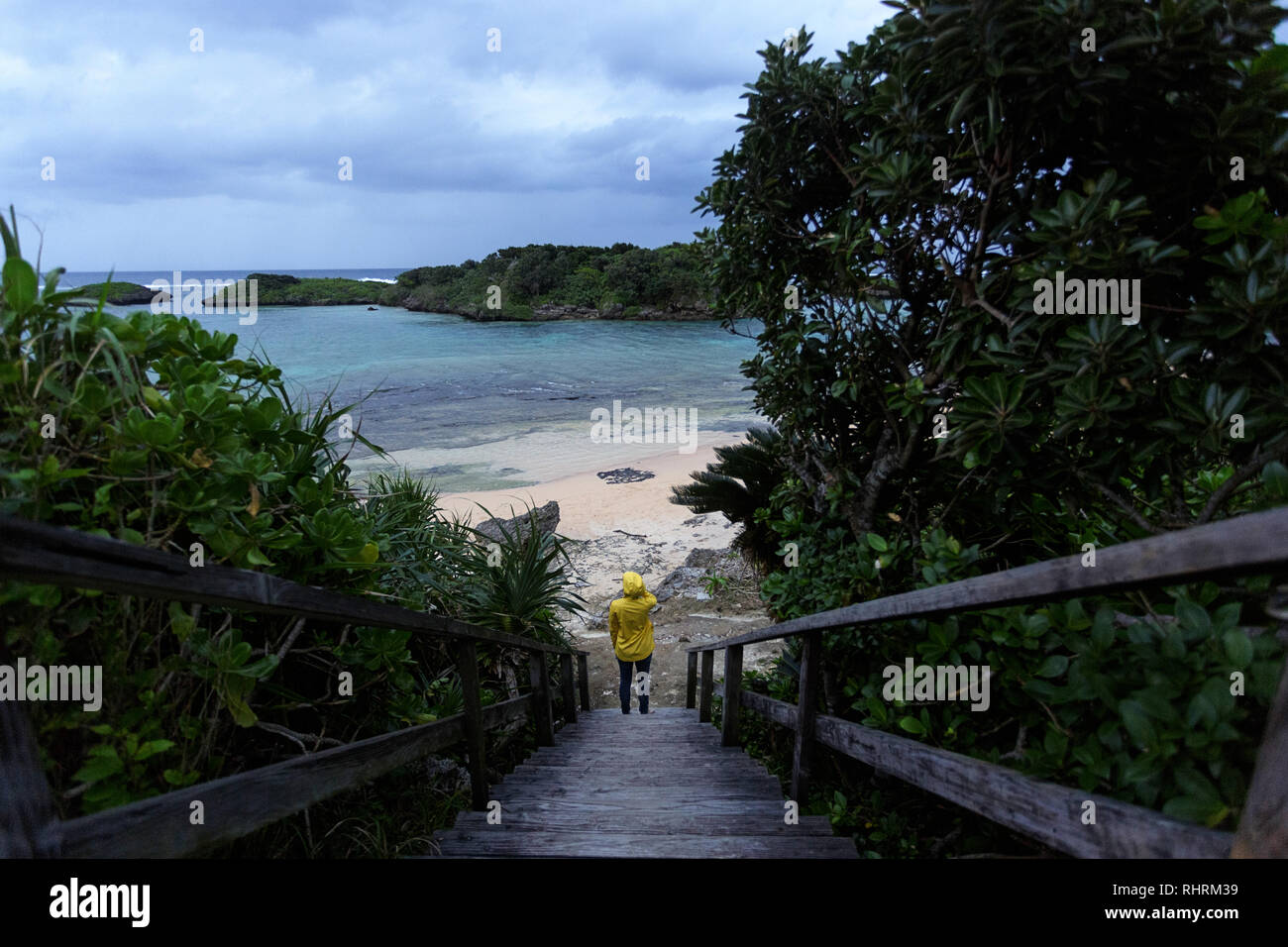 Femme en imperméable jaune debout sur trottoir de bois à la belle plage tropicale de sable par mauvais temps, le Japon Banque D'Images