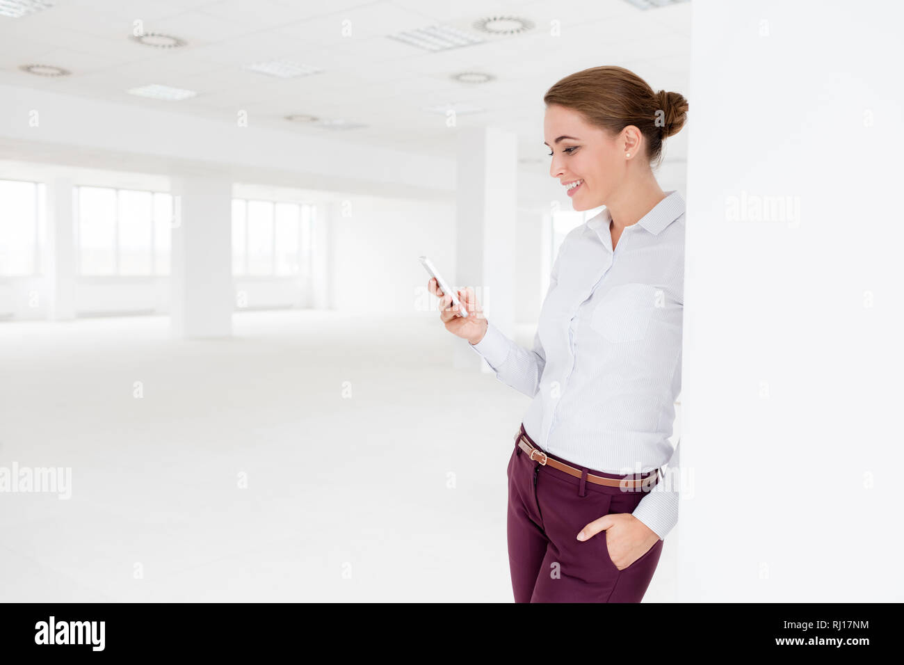 Smiling young businesswoman using smartphone pendant que debout à nouveau bureau vide Banque D'Images