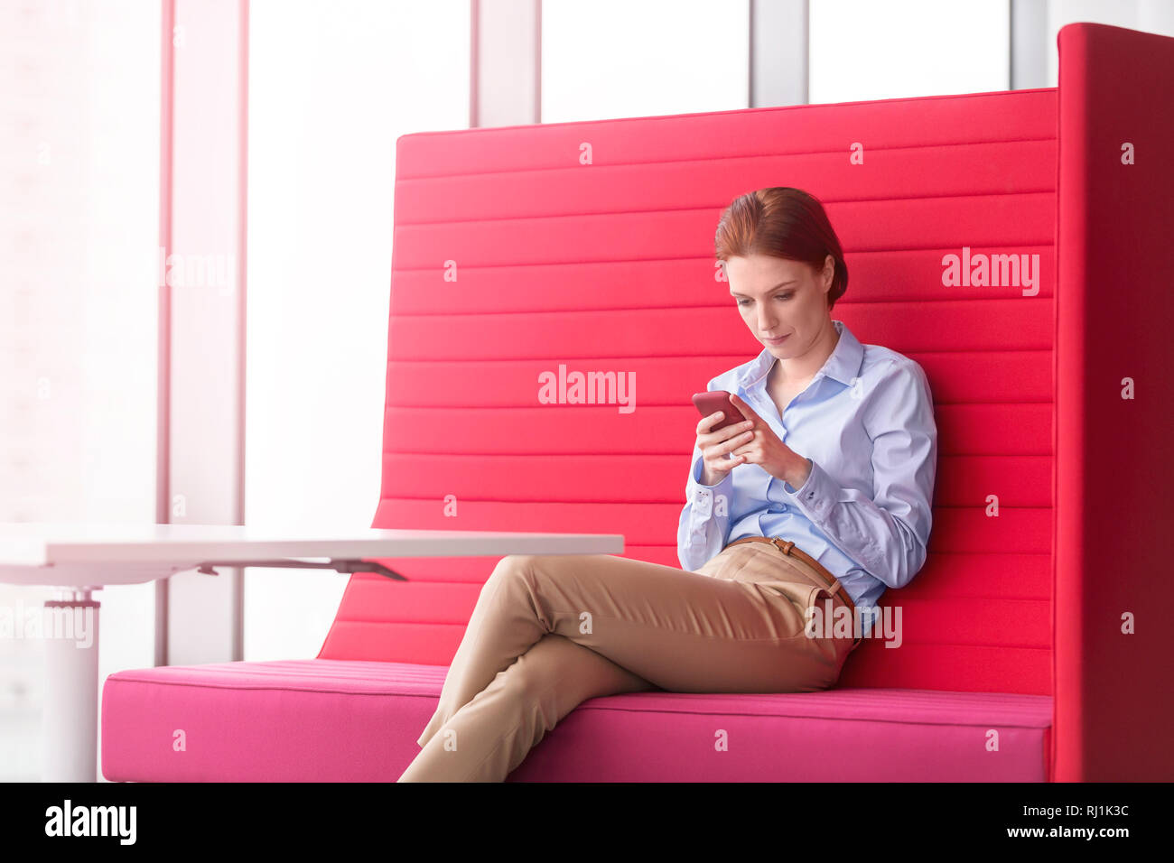 Young businesswoman messaging sur smartphone tout en étant assis sur le siège rouge at office Banque D'Images