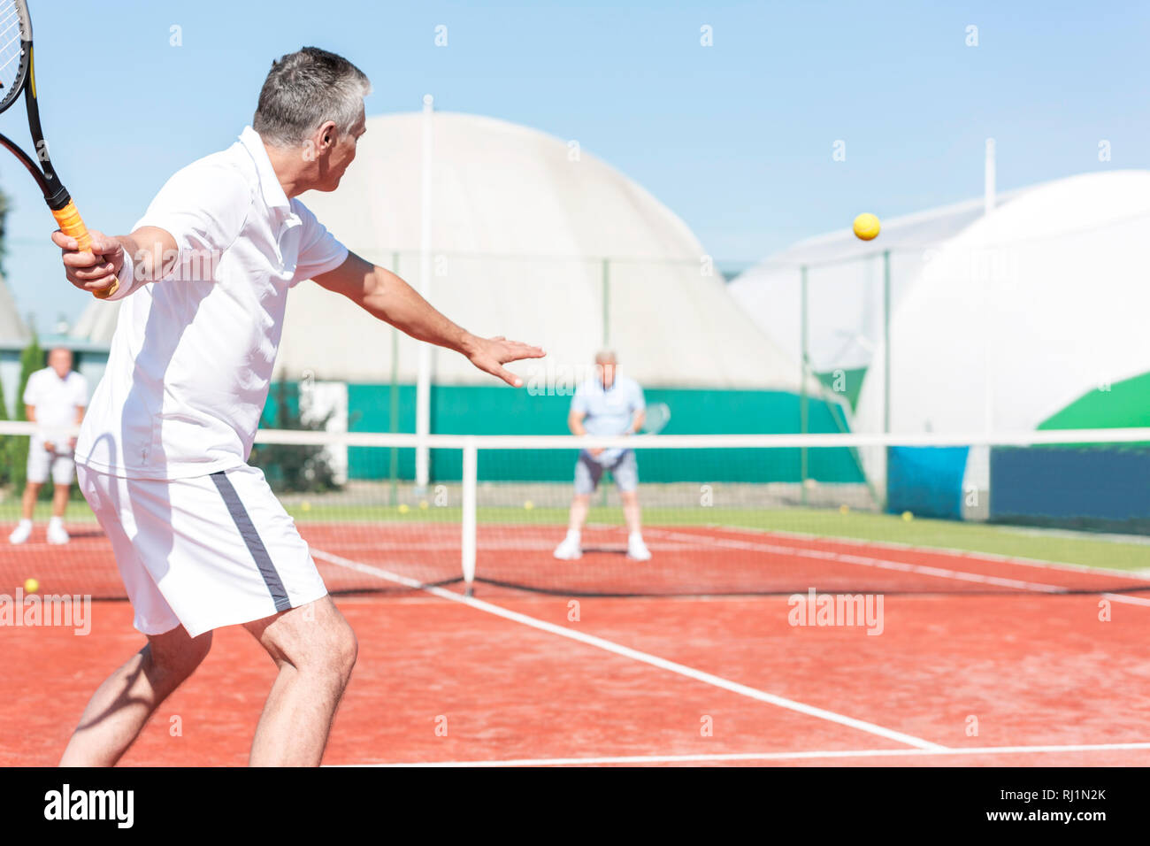 L'homme se balançant raquette de tennis tout en jouant match de double sur cour rouge pendant l'été, week-end Banque D'Images