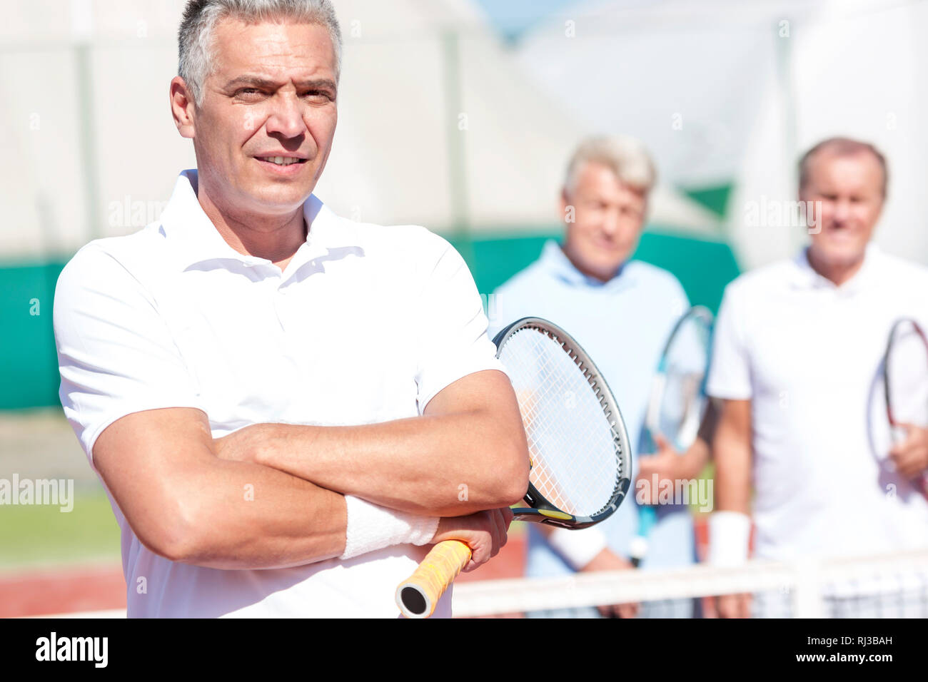 Portrait of mature man holding tennis racket en position debout avec les bras croisés contre des amis sur cour au cours de journée ensoleillée Banque D'Images