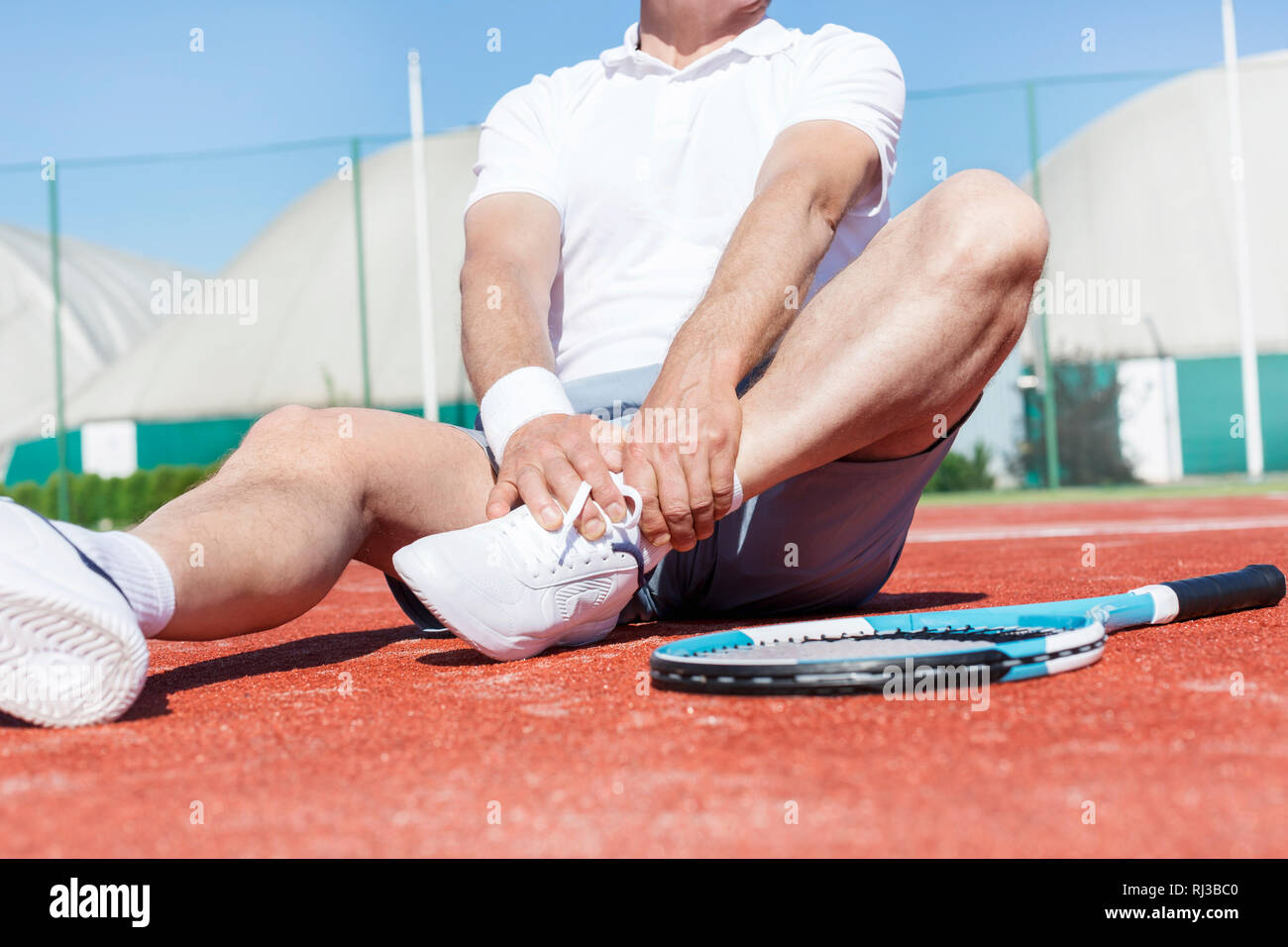 La section basse de l'homme mûr qui s'étend de la jambe en position assise sur le rouge de tennis en été Banque D'Images