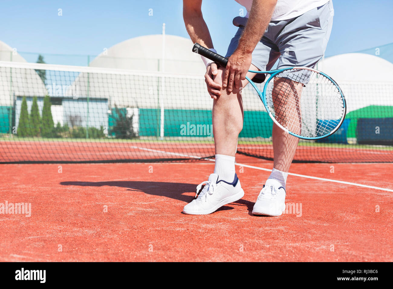La section basse de mature man holding tennis raquette tout en souffrant de douleur au genou sur rouge tennis durant l'été Banque D'Images