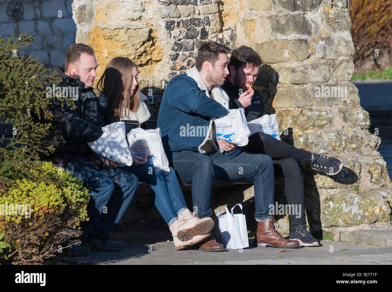 Groupe de jeunes gens assis à l'extérieur en hiver au Royaume-Uni La consommation de poisson et frites à partir de sacs en papier. Banque D'Images