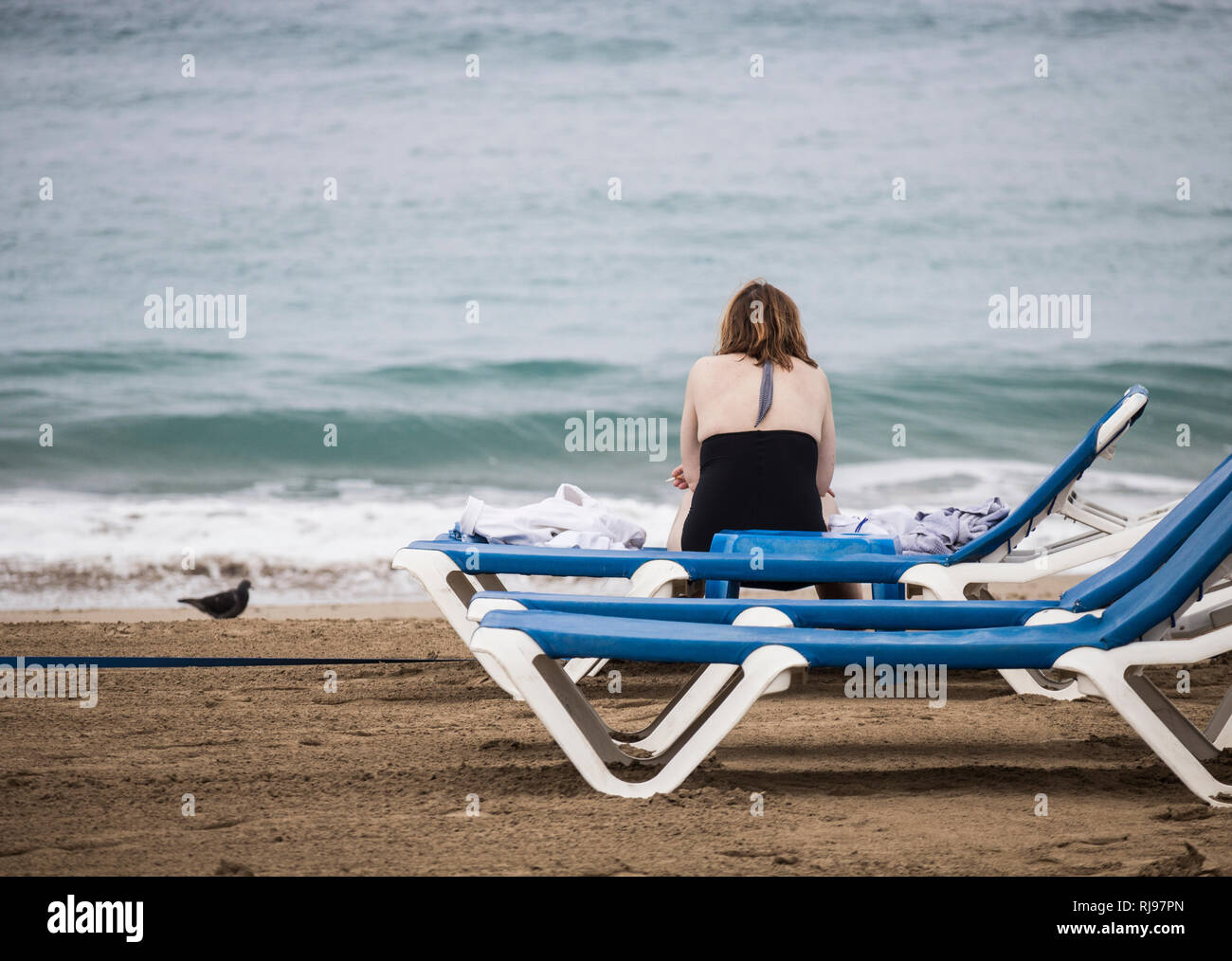 Vue arrière de la femme avec la cigarette à la main assis sur la plage transat sur un jour nuageux. Banque D'Images