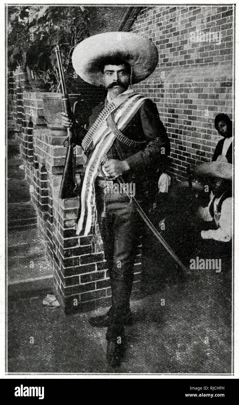 Emiliano Zapata se tient près d'un escalier en brique, dans un sombrero, tenant la garde de son épée et tenant un fusil en altitude, portant deux baudriers sur sa poitrine. Plusieurs hommes assis derrière lui dans l'arrière-plan. Banque D'Images