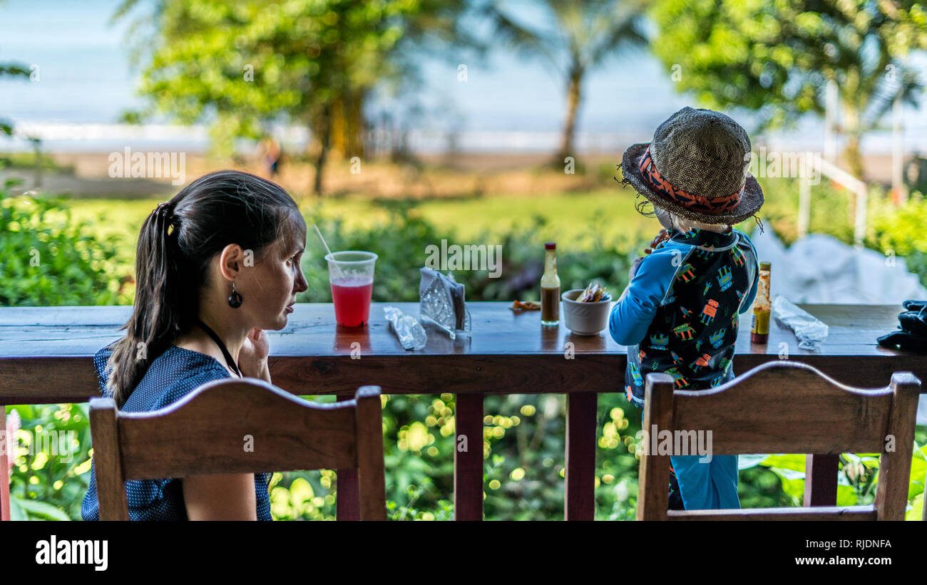 Une belle photo d'une mère et son fils ayant un repas et des boissons dans un diner à la belle plage de sable et vert de la baie Drake, Costa Rica. Banque D'Images