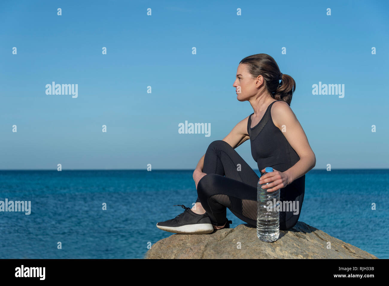 Grande sportive assis sur un rocher avec une bouteille en plastique d'eau de la mer au repos après l'entraînement et l'exercice. Banque D'Images